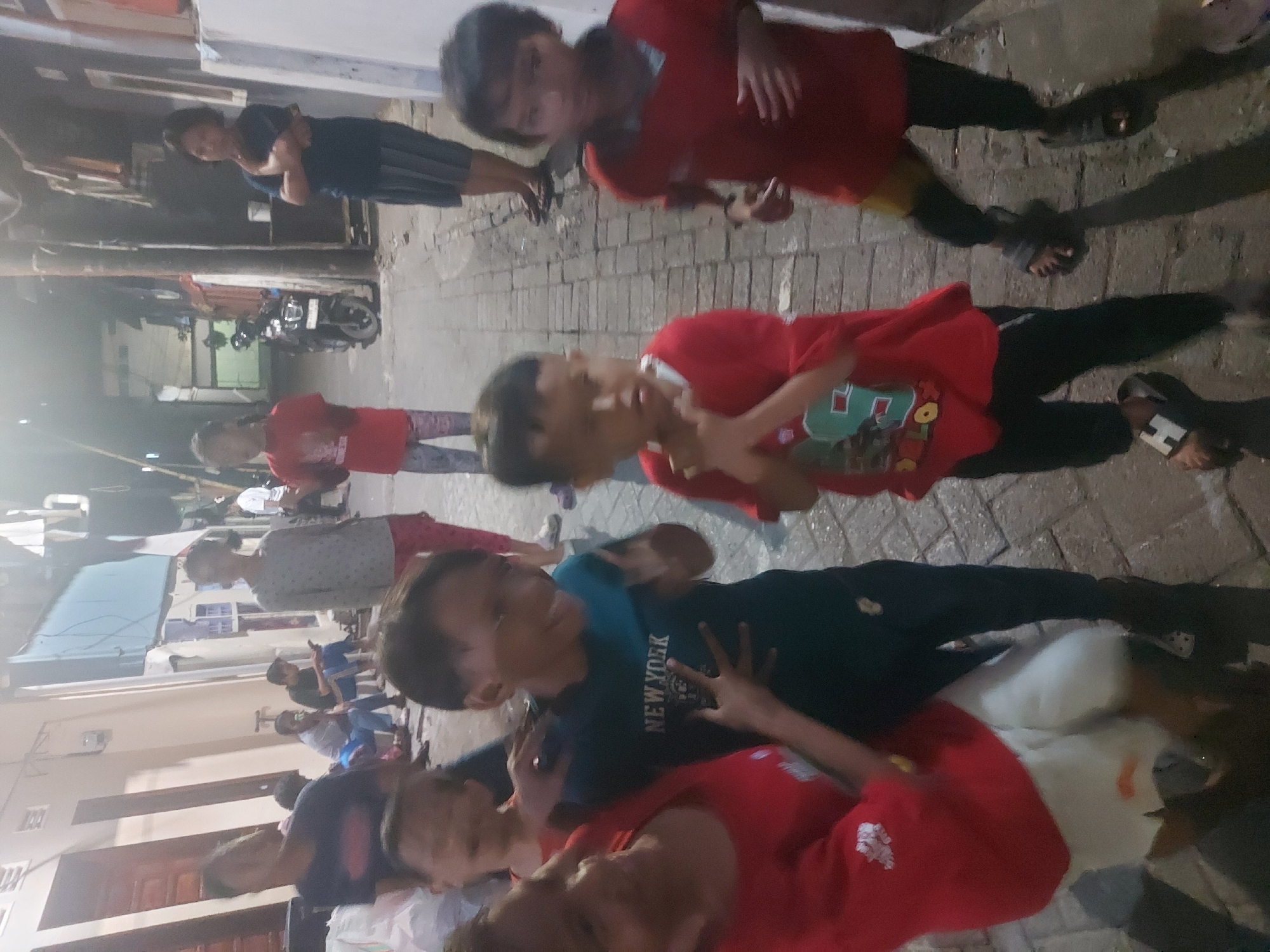 In Makassar, Indonesia, a group of approximately eight children, appearing to be of varying ages from early childhood to pre-adolescence, are gathered in a narrow, brick-paved alleyway. The lighting suggests it is dusk or evening. Several children are wearing red t-shirts and dark-colored pants or shorts, while others are in darker attire. One child in the foreground, wearing a dark blue t-shirt with "NEW YORK" printed on it, is actively engaged, with hands raised and a visible smile. Another child in a red t-shirt is also visible, looking towards the viewer. In the background, there are buildings with visible windows and doors, and a motorcycle is parked on the right side of the alley. The overall atmosphere appears casual and communal, with the children interacting amongst themselves in a typical urban residential setting.