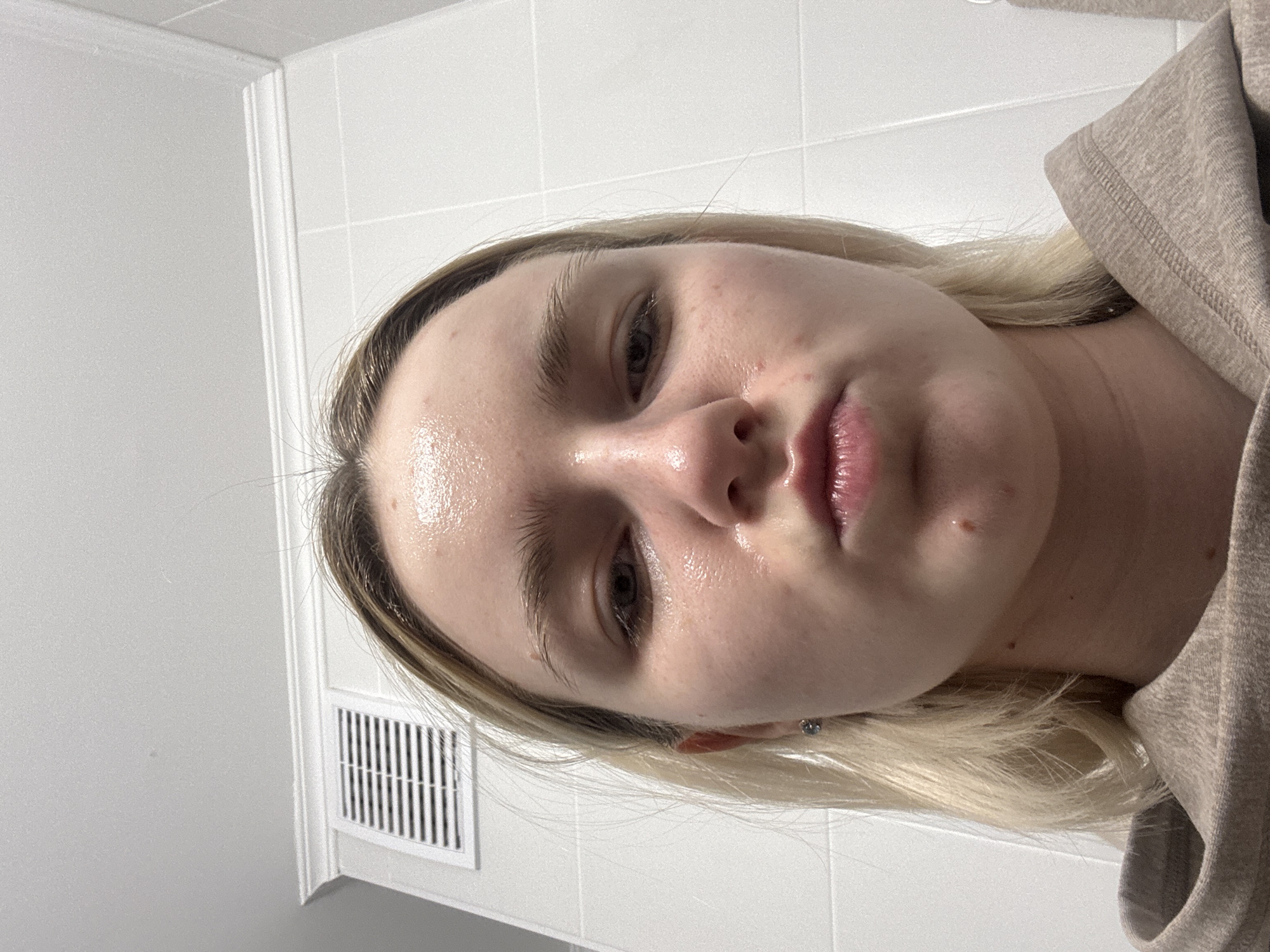 A close-up selfie shows a young woman with fair skin and light blonde hair, looking directly at the camera. Her face has a noticeable sheen, particularly on the forehead, nose, and chin, suggesting oily skin or recently applied moisturizer. Minor blemishes are visible on her cheeks and chin. She has dark eyes and a neutral, perhaps slightly pensive, expression. She is wearing a light brown or beige hooded sweatshirt, the fabric of which is visible on her right side. The background consists of white tiled walls, typical of a bathroom or utilitarian indoor space. A white rectangular air vent or return grill is visible on the wall to the left of the woman. The lighting is bright and even, consistent with an indoor, artificially lit environment. No text is visible in the image.