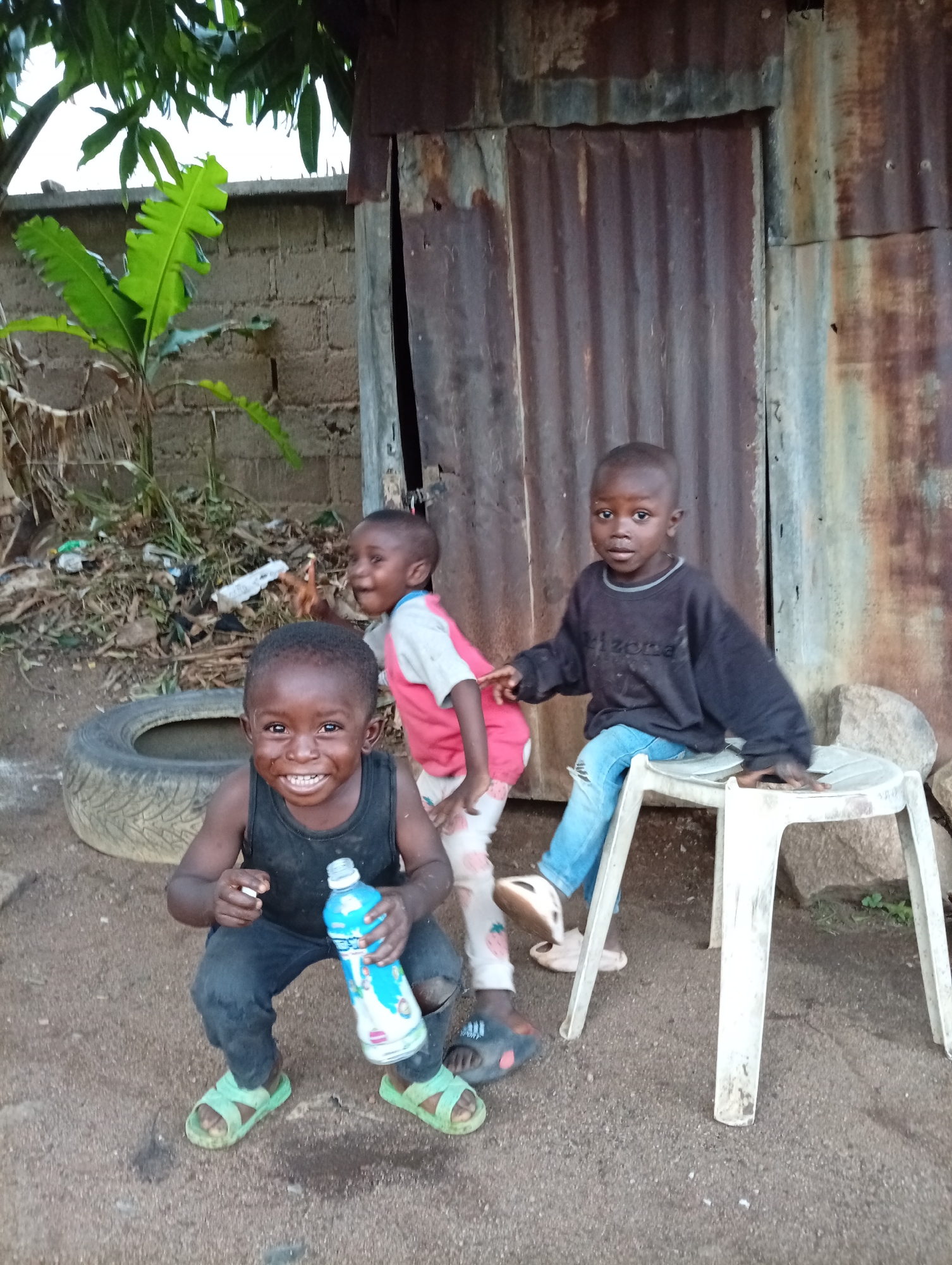 Three young children are located in Bukuru, Nigeria, situated in a yard near a rusted, corrugated metal structure that appears to be a shed or small outbuilding. In the foreground, a child with dark skin wears a black tank top, dark pants, and green sandals, holding a blue water bottle and smiling broadly. Behind him, another child in a red shirt and patterned pants has a joyful expression. To the right, a third child sits on a white plastic chair wearing a black sweatshirt with blue jeans and slippers.
A tire lies on the ground near the children and the metal building. In the background is a cinderblock wall overgrown with vegetation. The ground is dirt, suggesting a rural or impoverished setting. The overall image depicts poverty and a lack of sanitation, with trash visible in the background.