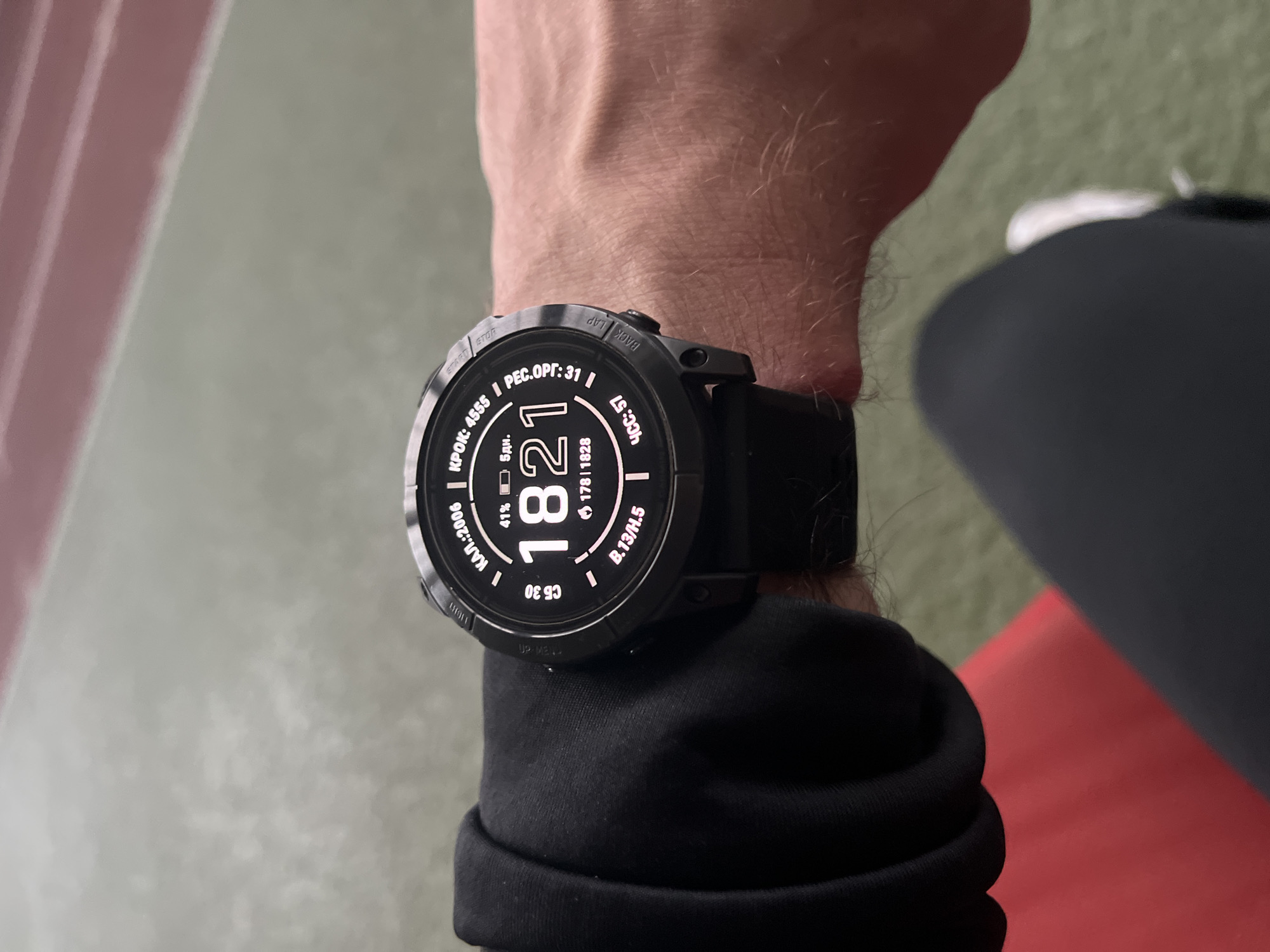A close-up shot shows a person's wrist with a large, black smartwatch. The watch display shows various metrics including time (18:21), battery percentage (41%), steps (4555), and heart rate (57). The watch has multiple buttons and a black wristband. The person is wearing a black long-sleeved shirt or jacket. The background consists of blurred surfaces with a possible red stripe visible.
