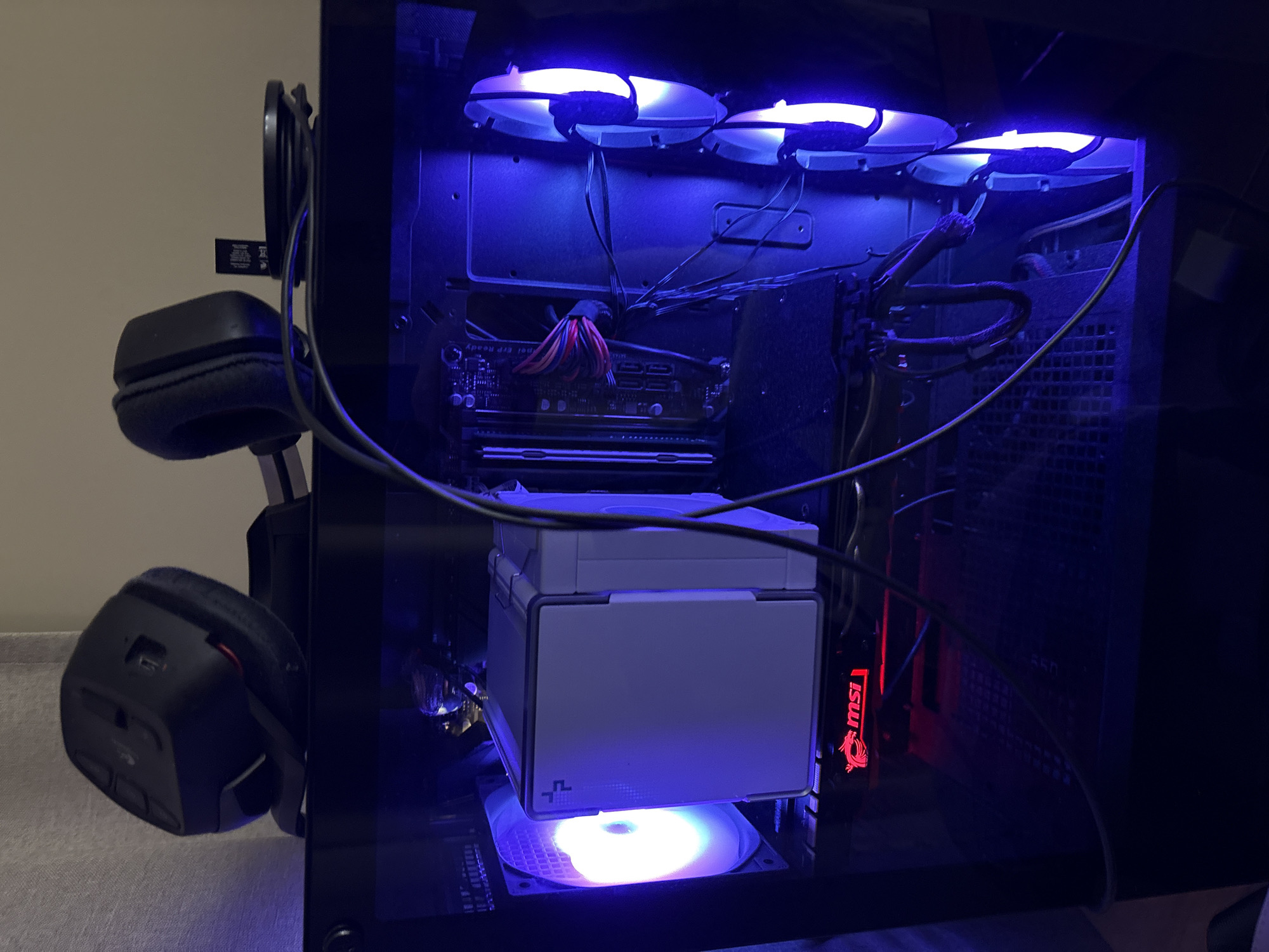 The image displays the illuminated interior of a desktop computer tower, viewed through a transparent side panel, in a dimly lit indoor setting. Bright blue LED lighting emanates from three cooling fans mounted at the top of the case, and from other internal components. A prominent, white and grey CPU cooler is situated centrally, beneath which a fan emits a softer white or light blue glow, with a partially visible "DP" or "DeepCool" logo on the cooler.
To the right, an MSI graphics card is visible, featuring a distinct red illuminated "msi" logo. Various internal cables are seen, some neatly bundled, others spanning across the components. A section of a motherboard is also visible towards the top, with faint, unreadable text like "Xpea".
On the left exterior side of the PC case, a pair of black, over-ear headphones hang from a hook or bracket. Their cable trails into the computer case. A small, black dongle-like device with indecipherable text is also attached near the headphones. The overall setup suggests an active computing or gaming station within a home environment in Sofia, Bulgaria.