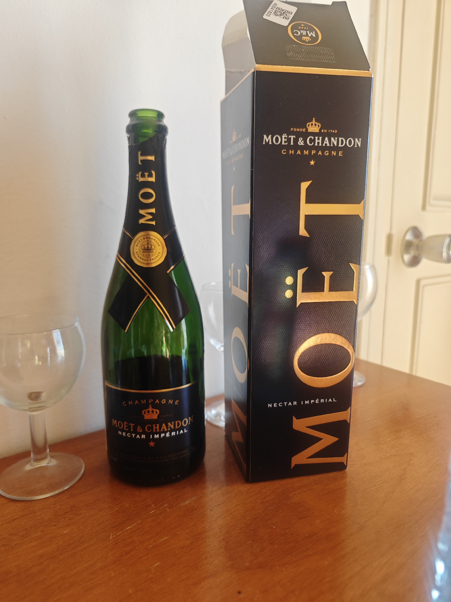 The image shows a Moët & Chandon champagne bottle and its presentation box on a wooden table. The bottle is green glass with a black and gold label. The presentation box is black with gold lettering and a flap. There are two clear champagne flutes next to the bottle. The background consists of a white wall and a wooden door with a doorknob. The scene appears to be an indoor setting, possibly a home or a restaurant, and the location in Hiboun, Tunisia, is not discernible from the image. There are no people or animals present in the image, and no actions or interactions are depicted.