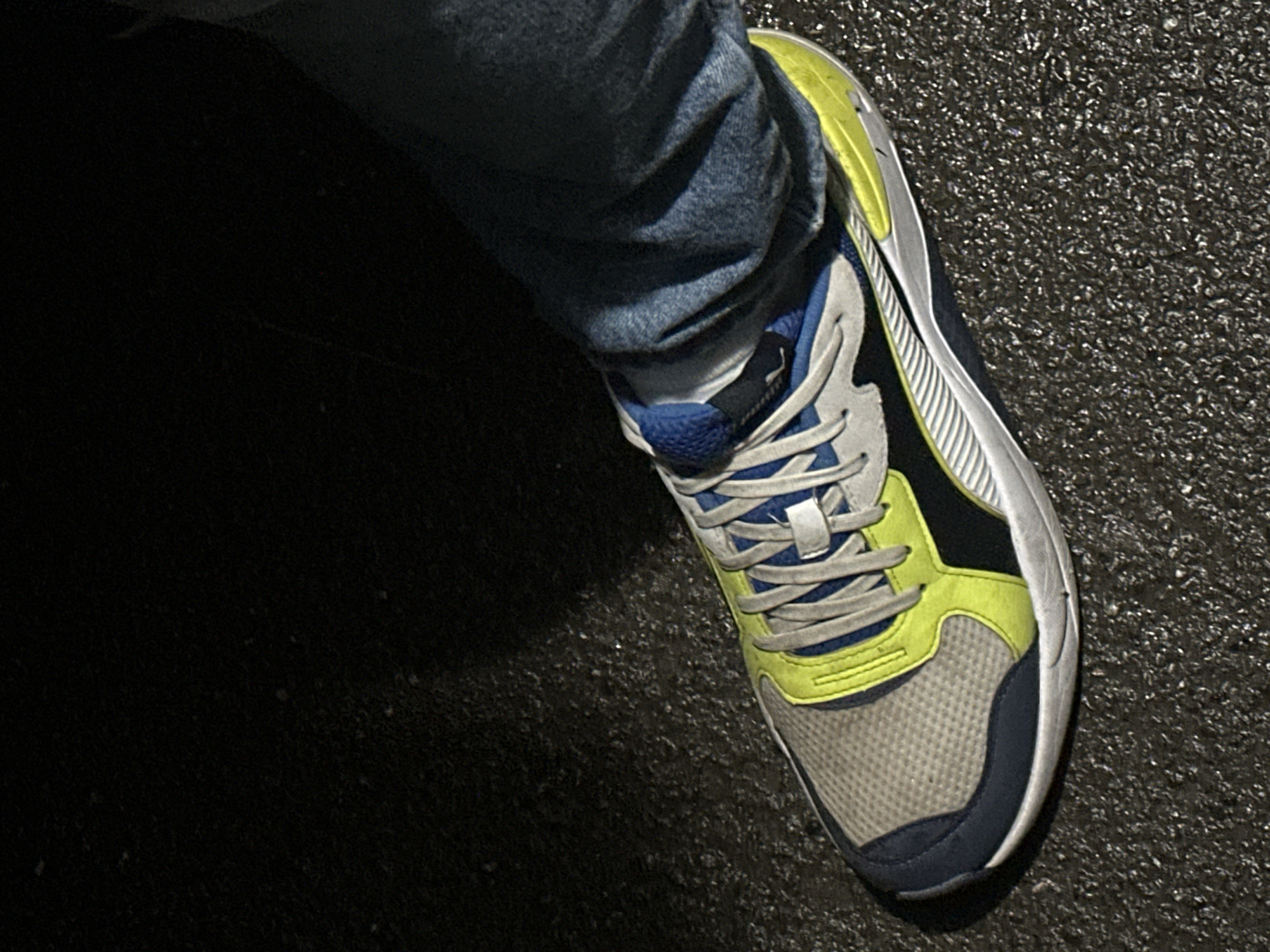 The image shows a close-up, low-angle view of a person's foot wearing a white, blue, black, and neon yellow athletic shoe. The shoe is positioned on a dark, wet, asphalt surface, possibly a road or sidewalk. The person is also wearing blue denim jeans. The lighting suggests it might be nighttime or a dimly lit indoor space, with a spotlight illuminating the shoe. There is no identifiable context for the location being Togliatti, Russia; this information is not present in the visual data. The image is a snapshot of footwear and apparel in an urban or semi-urban environment.