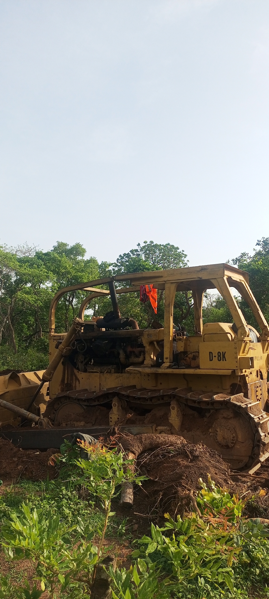 The scene depicts a yellow Caterpillar D-8K bulldozer situated in a vegetated area, possibly on the outskirts of Otunade, Nigeria. The bulldozer appears used, with mud and debris covering its tracks and undercarriage. An orange cloth is draped inside the roll cage of the machine. The surrounding environment consists of green foliage, including bushes and trees, suggesting a rural or undeveloped setting. The sky is a pale blue, indicating daylight. No people are visible in the image. The bulldozer appears to have been operating recently, given the disturbed earth around it.