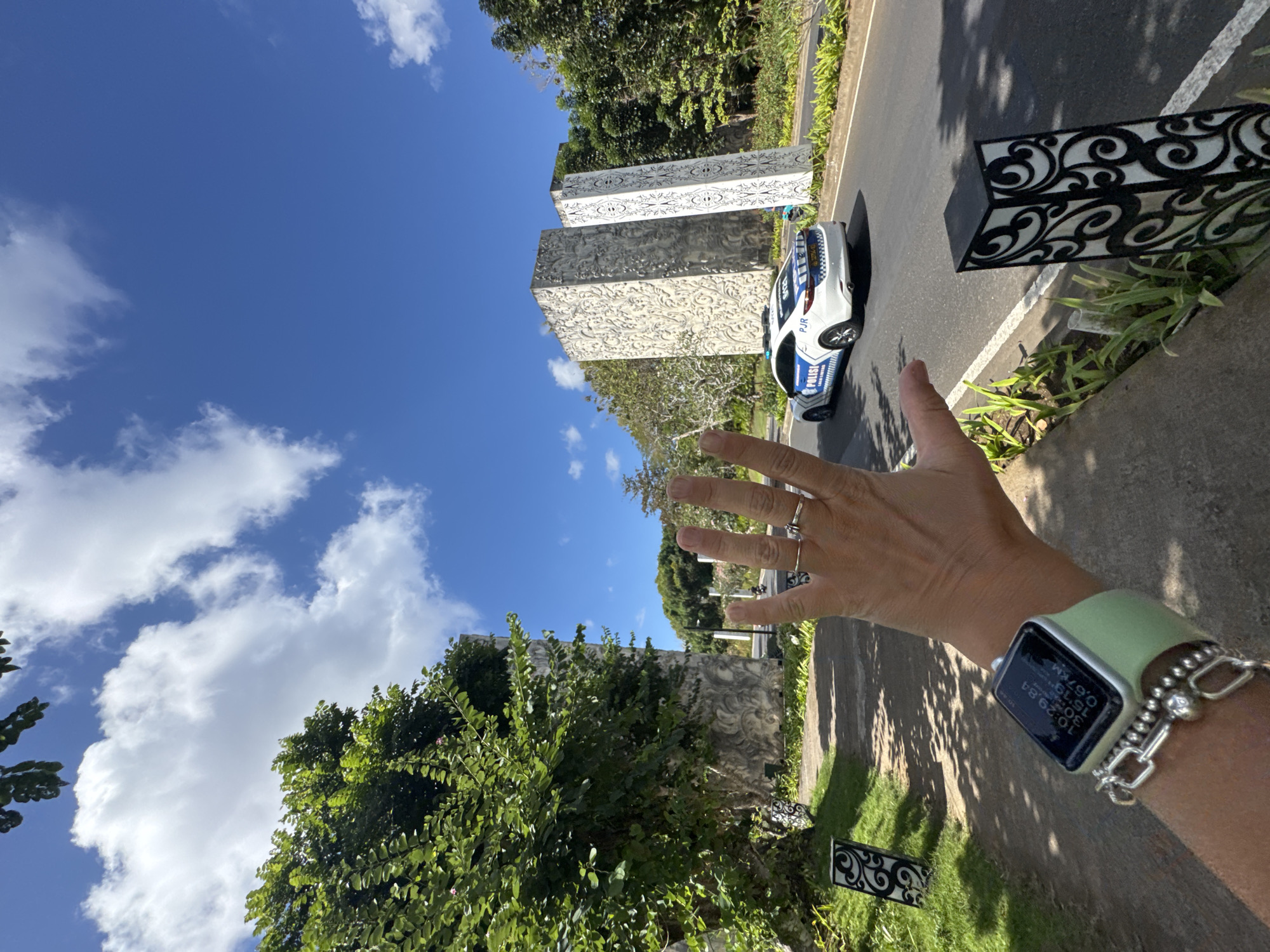 An extreme wide-angle, eye-level shot captures a person's extended left hand and wrist in the foreground, with a police car and large stone pillars in the background, set against a bright blue sky with scattered clouds. The hand, adorned with two rings, is positioned to obscure the middle and ring fingers. A smartwatch with a light green band is visible on the wrist, displaying a time of 10:29:54, distance of 0.67 km, and possibly other metrics. A bracelet with silver beads and a chunky silver chain is also worn on the wrist. The police car, a white sedan with blue and white markings that read "POLISI" and "PJR", is driving on a paved road that curves towards the right. The road is bordered by a manicured lawn and decorative black posts with white scrollwork. To the left of the road, a large, textured stone pillar with intricate carvings stands prominently, partially obscured by lush green foliage. Another similar, but taller, stone pillar is visible further down the road. The environment appears to be tropical, with abundant greenery and tall trees. The presence of the police car suggests a location within Kuta Selatan, Indonesia, possibly an entrance or checkpoint.