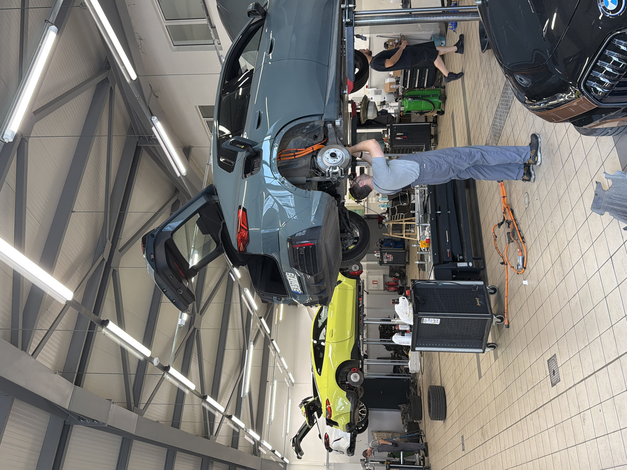 The image depicts a car repair shop in Burgas, Bulgaria, specifically the BMW service center. Two car lifts are occupied by vehicles. The first lift holds a gray BMW with its trunk open, displaying orange electrical cables near the rear wheel well. A person in gray overalls is working on the underside of this vehicle. To the left, a yellow BMW is on a second lift, also with its trunk open, and a white BMW is parked next to it. Another person in a black t-shirt and shorts is visible in the background, walking away from the viewer. The environment is a brightly lit indoor workshop with a tiled floor, metal support beams, and fluorescent lighting. Various tools, equipment, and car parts are scattered around the workspace.