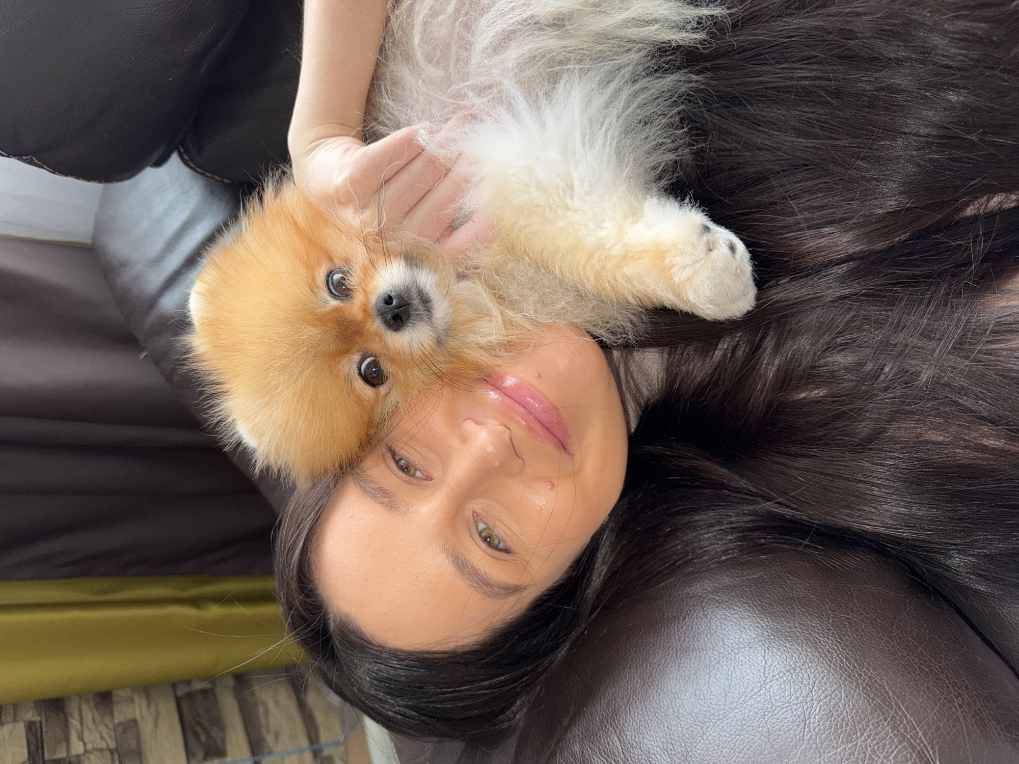The image displays a close-up, top-down view of a woman and a small, fluffy dog, likely a Pomeranian, in an affectionate pose. Both subjects are lying down, presumably on a dark brown leather couch or armchair, which forms the primary background.
The woman has long, straight dark brown hair fanned out around her head. She possesses fair skin, light-colored eyes (appearing greenish-brown), and soft pink, glossy lips. Her expression is serene and relaxed as she looks towards the camera. Her left arm is visible, gently supporting or caressing the dog.
The small dog has golden-orange fur around its face and a lighter, whiter fur on its body, suggesting a mix of colors. It has dark, expressive eyes and a small, black nose, and is looking directly into the camera with a calm demeanor. The dog's body is nestled against the woman's chest or neck area.
The scene conveys a moment of warmth, companionship, and relaxation between the woman and her pet. The lighting appears soft and natural, indicative of an indoor setting during the daytime. No specific time of day or external weather conditions are discernible. No visible text is present in the media file. This tender moment takes place in Khimki, Russia.