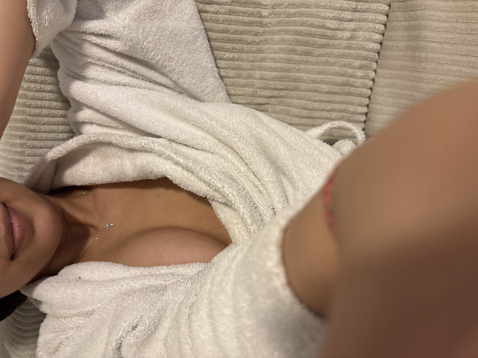 The image captures a close-up, high-angle view of a person lying down, predominantly dressed in a white, fluffy bathrobe. The subject's lower face, featuring a slight smile on their lips, is visible on the left side of the frame. A delicate silver-colored necklace with a small cross pendant adorns their neck and collarbone. The bathrobe is open, revealing a portion of their chest.
The material of the bathrobe appears to be a soft, terry-cloth texture. In the background, behind the person and the bathrobe, lies a light beige or greyish-brown surface, likely a cushioned item like a pillow, blanket, or couch upholstery, distinguished by prominent horizontal ribbed patterns. A blurred, light-skinned limb is partially visible in the right foreground, suggesting the image is a selfie. The scene is illuminated by indoor, ambient light, creating a relaxed atmosphere. No specific text is visible in the image, nor are there any visual cues to pinpoint the exact time of day or specific outdoor location beyond an interior setting. The person's expression indicates a state of relaxation or contentment.