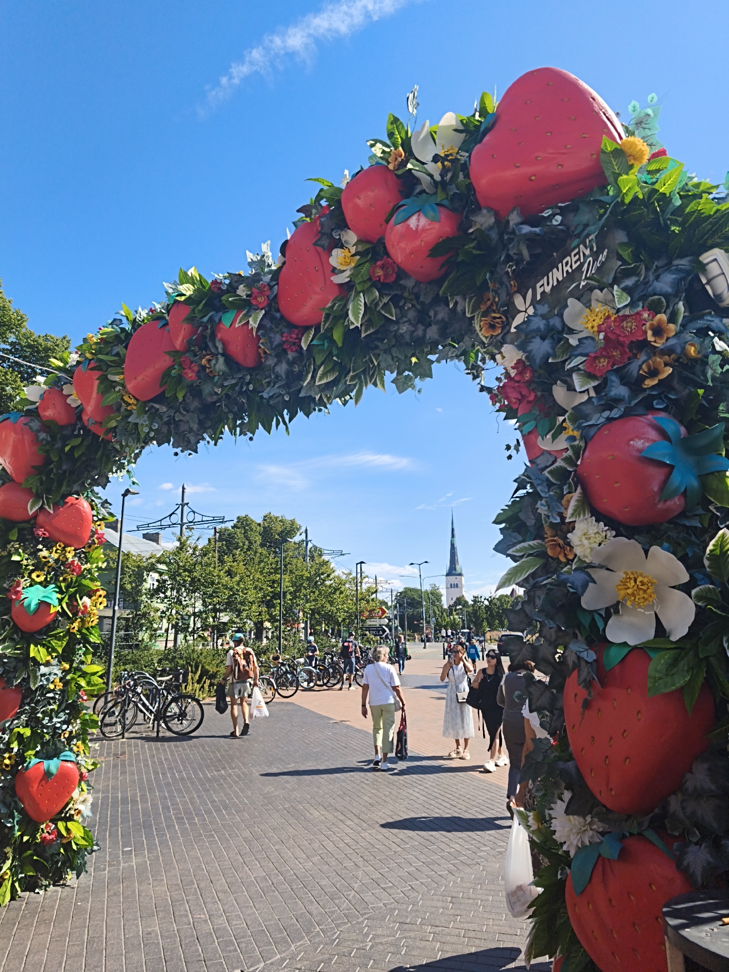 The image depicts a view through a large, festive archway adorned with oversized artificial strawberries, green foliage, and flowers. The archway is situated on a paved pedestrian walkway in Tallinn, Estonia, likely during a summer event or festival. A tall, slender church spire is visible in the distance, characteristic of Tallinn's Old Town.
In the foreground, several people are walking on the brick-patterned pathway. To the left, a rack of bicycles is parked. The sky above is clear blue with a few wispy clouds. The overall impression is a bright, sunny day in a bustling urban environment transformed by a whimsical, fruit-themed decoration.
The archway itself is a prominent feature, acting as a frame for the scene beyond. The strawberries are bright red and appear to be made of a smooth, molded material. They are interspersed with artificial green leaves and smaller decorative flowers in shades of white, red, and yellow. A small sign with the text "FUNRENT Deco" is visible on the archway.
The pedestrian walkway is made of grey brick pavers laid in a herringbone pattern. Several people are seen walking along it, some carrying shopping bags. Their attire suggests warm weather. On the left side of the walkway, near the bicycles, a person with a backpack is walking away from the camera.
In the mid-ground, trees line the walkway, and more people and bicycles are visible. The church spire in the distance provides a strong architectural landmark. The scene suggests a lively and pleasant atmosphere.