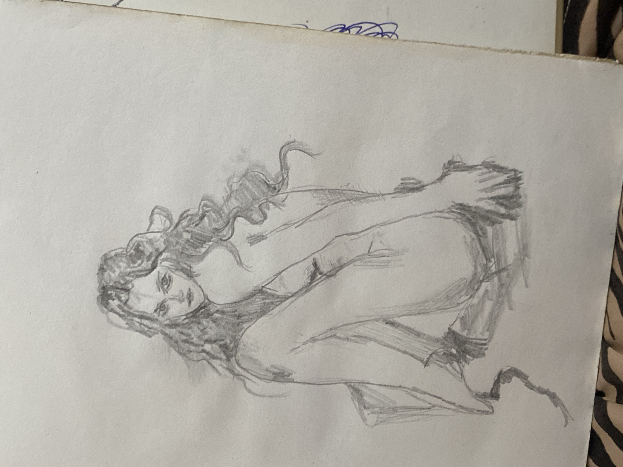 This is a pencil sketch of a person in a dynamic pose. The subject appears to be female, with long, wavy hair. She is positioned on her side, with her legs bent and her arms wrapped around herself, suggesting a sense of enclosure or self-protection. Her facial expression is somber and her gaze is directed towards the viewer, conveying a feeling of introspection or perhaps distress. The shading and linework are somewhat loose and gestural, giving the drawing a raw and expressive quality. The sketch is on a piece of paper that shows some wear and tear at the edges. There is some blue ink scribbling visible at the top edge of the paper, but no other discernible text. The overall mood of the drawing is melancholic and intimate. The context provided indicates the location is Zhmerynka, Ukraine, but there are no visual cues within the sketch to support or contradict this.