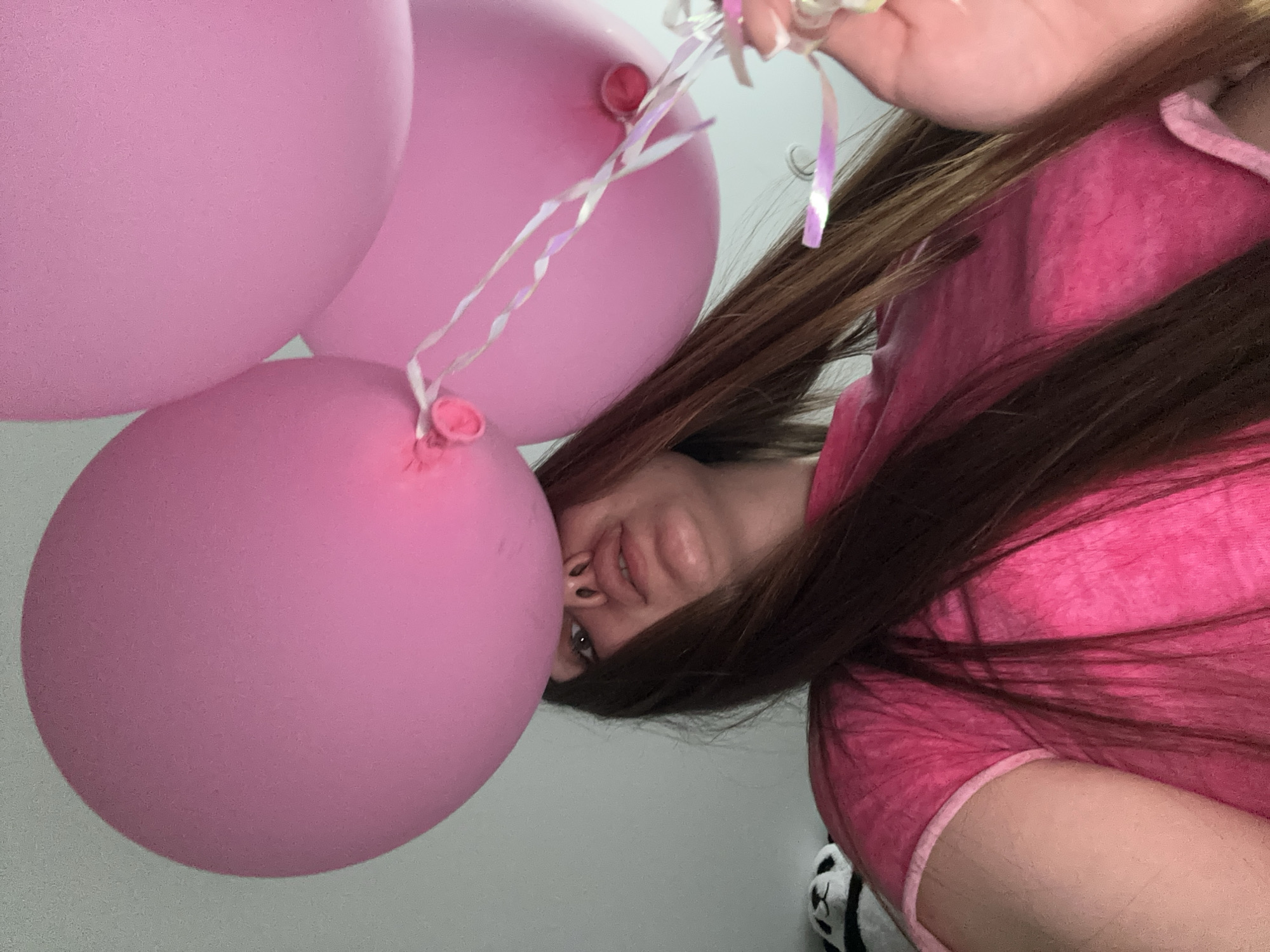 A woman with long brown hair and wearing a pink shirt is holding three pink balloons. The photo appears to be taken from a low angle looking up at the woman, and the balloons are positioned above her. The background is a plain white wall. A panda plush can be seen near the bottom-right corner. It is not possible to ascertain the location of the photo, and specifically the city of Chaikovsky, Russia, based on the visual information provided.