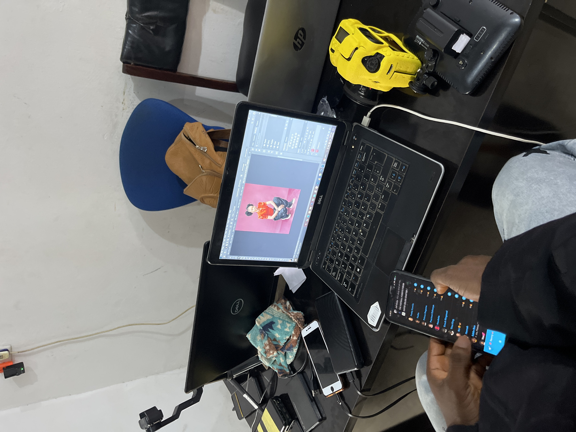 This media file shows a top-down view of a desk cluttered with electronic devices, a tan jacket, and a blue chair. Two laptops are open, one displaying photo editing software with an image of a person in an orange shirt and black pants. A black smartphone is being held by a person whose hands and arm are visible in the lower right corner. The phone screen shows a list of names and numbers, possibly a contact list or chat application. Other objects on the desk include a yellow toy resembling a police car or drone, a black electronic device, and various cables and accessories. The background appears to be a wall with some light fixtures. The setting suggests a workspace or a personal area where someone is engaged with their electronics. The overall impression is one of a busy, tech-oriented environment. Visible text includes "DELL," "hp," "ATITUD," and some text on the smartphone screen that appears to be names and possibly chat-related content. The location context is Gura Top, Nigeria.