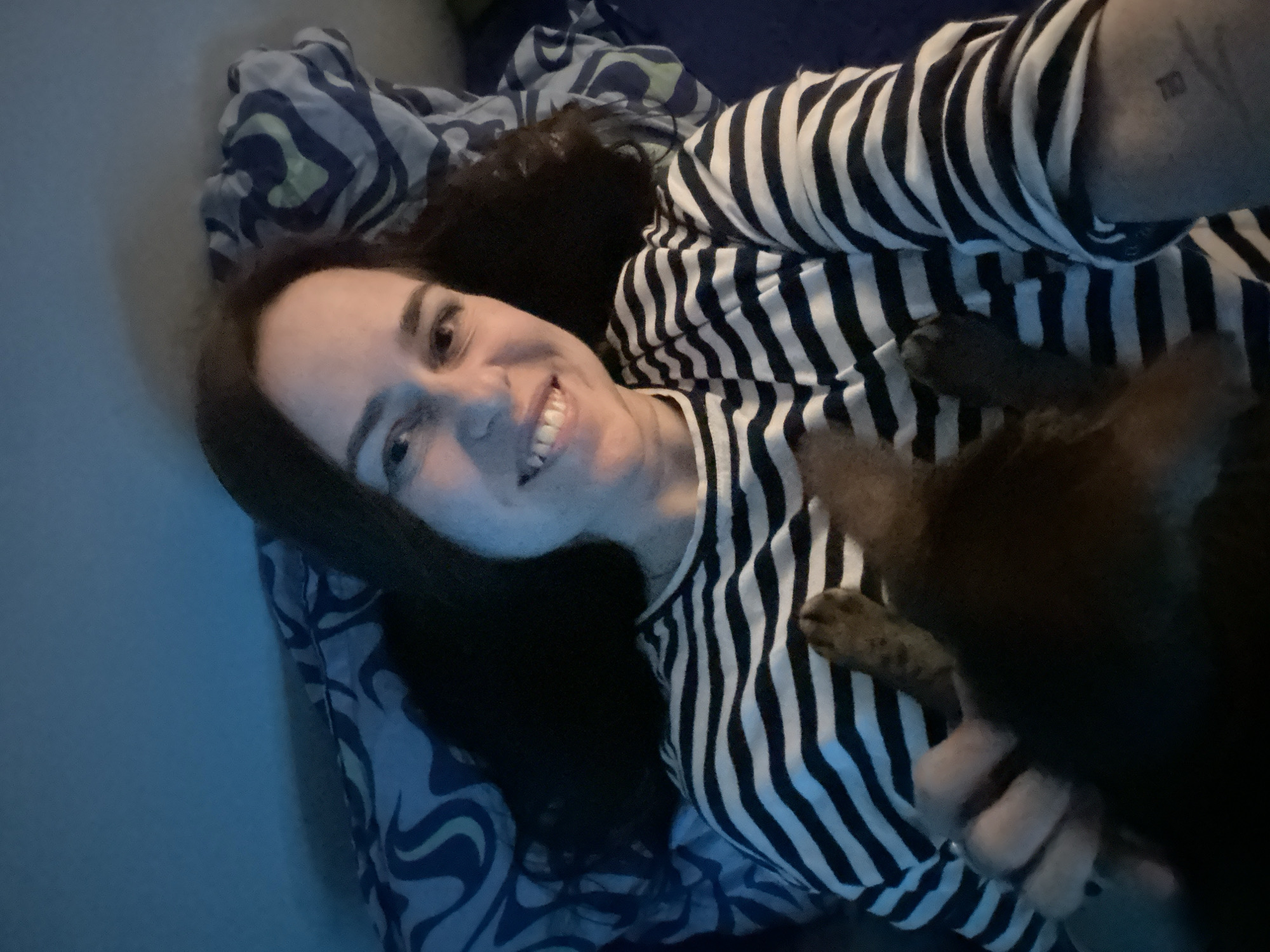 A woman with dark hair is lying on a bed with a patterned blue and white sheet. She is wearing a black and white striped shirt and smiling. A dark-colored cat is partially visible in the foreground, with its paws resting on the woman's shirt. There is a tattoo visible on the woman's arm. The image's lighting is somewhat dim.
There is no information available to indicate that this image was taken in Moscow, Russia.