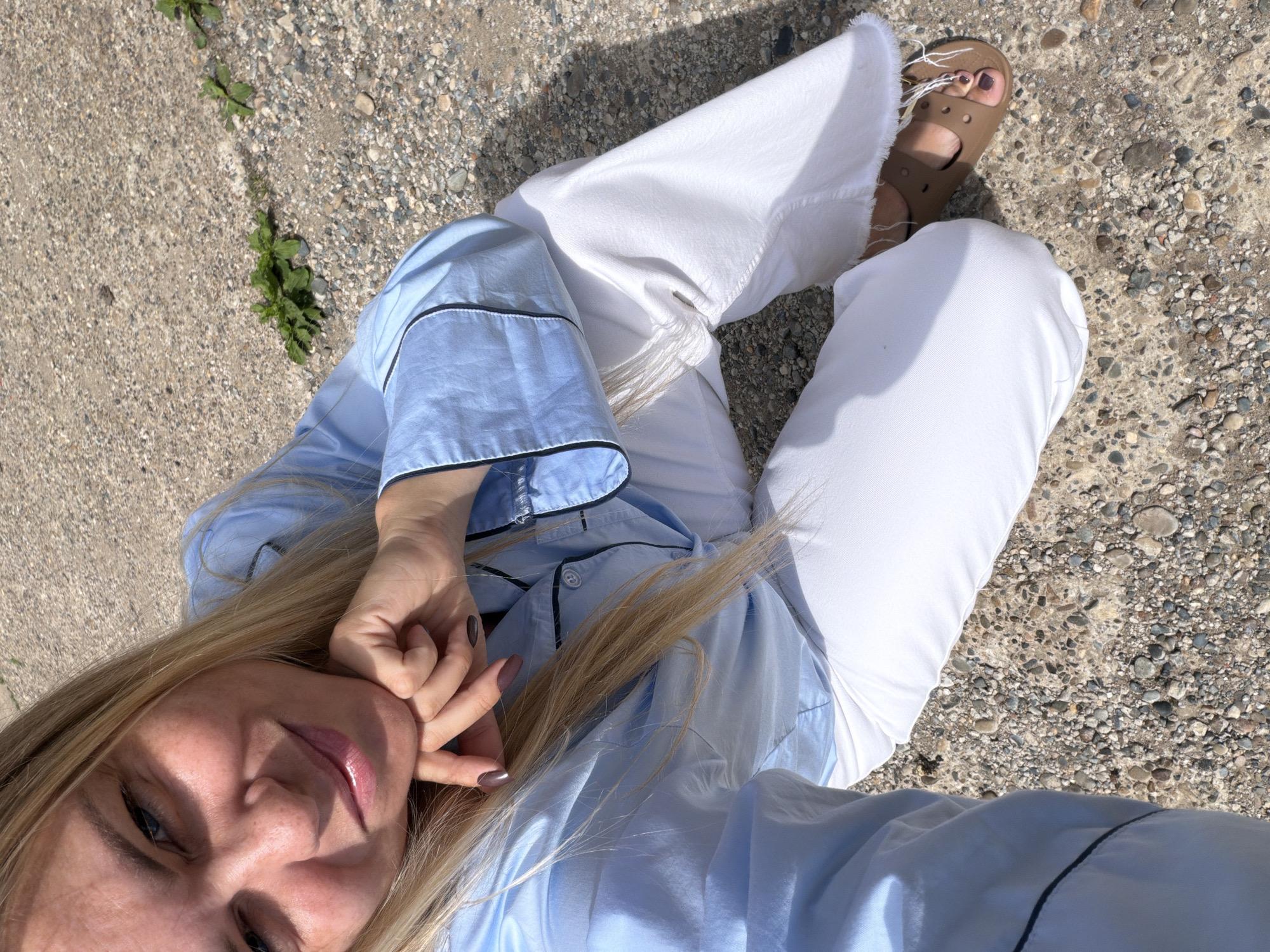 A young woman with blonde hair is kneeling on a gravelly surface, looking into the camera with a smile. She is wearing a light blue pajama top with black trim and white wide-leg jeans with frayed hems. Her hands are resting on her chin, and her nails are painted a dark color. She is wearing tan slip-on sandals. The background consists of the same gravelly ground and some sparse green plants. The lighting suggests it is daytime with bright sunlight. The location is not identifiable as Votkinsk, Russia, from the image alone.