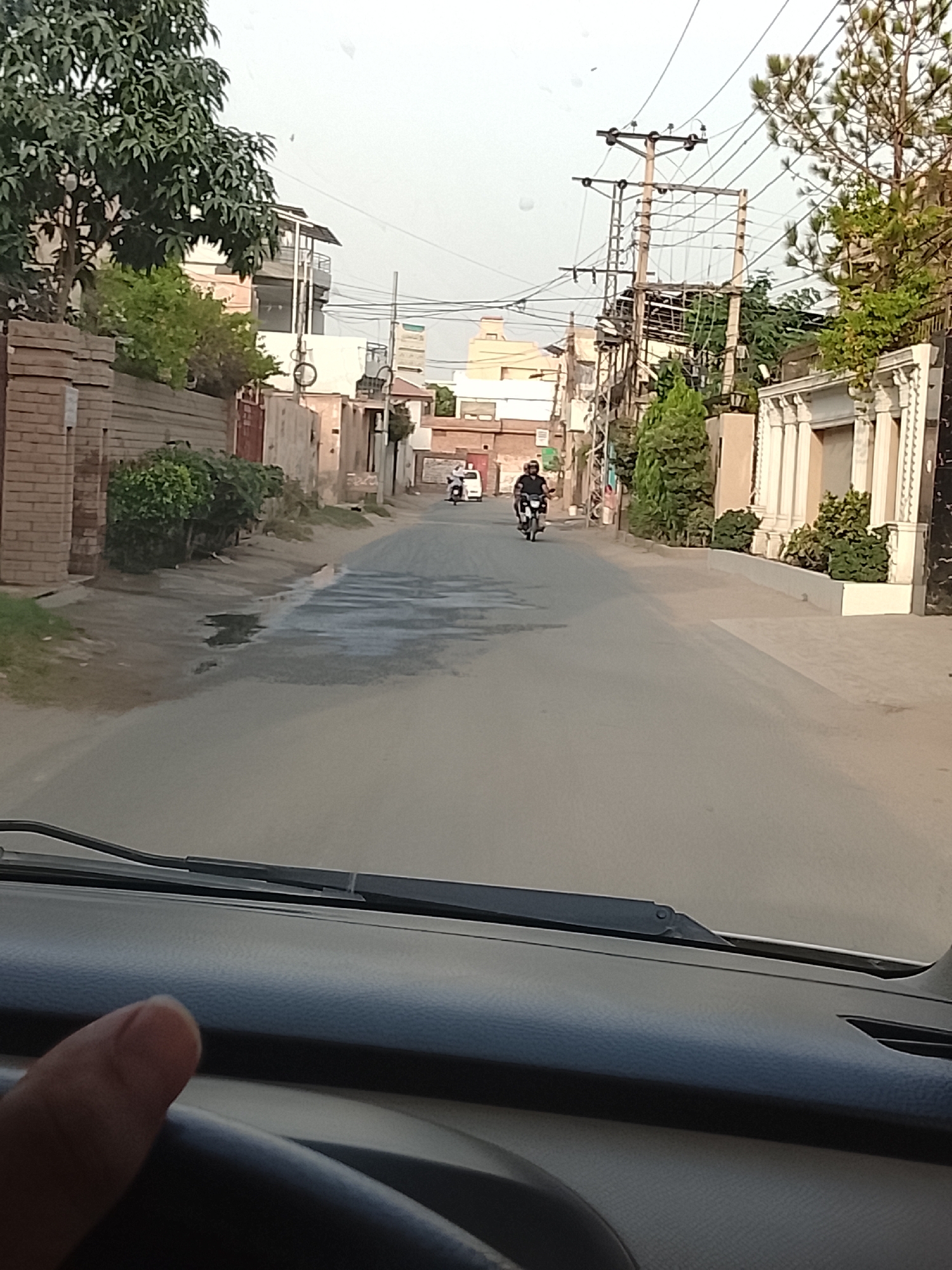 The image, seemingly taken from inside a vehicle moving along a road in Gulgasht Colony, Pakistan, depicts a street scene.
The street is flanked by houses and boundary walls on both sides. Trees and foliage are visible. There are some puddles on the left side of the road. An electrical pole with multiple wires runs along the right side.
Two motorcycles are on the road. One is traveling in the same direction as the camera, and is closer to the camera. A second motorcycle is further down the road, traveling in the opposite direction.
The dashboard and windshield of a vehicle are visible in the foreground. A finger is partially visible on the left side of the image.