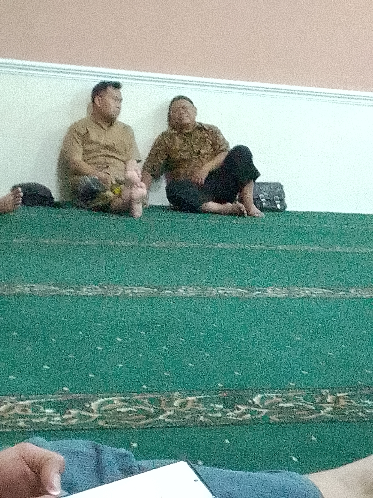 Two adult males are seated on a raised green carpeted platform. The male on the left is wearing a tan short-sleeved shirt and is holding a pink stuffed toy. The male on the right is wearing a brown and tan patterned short-sleeved shirt and dark pants. A black bag is to his right. The background consists of a white wall with a beige ceiling. The scene appears to be indoors, possibly a place of worship or community gathering in Kertajaya, Indonesia. A person's hand is visible in the lower portion of the image, holding a white tablet.