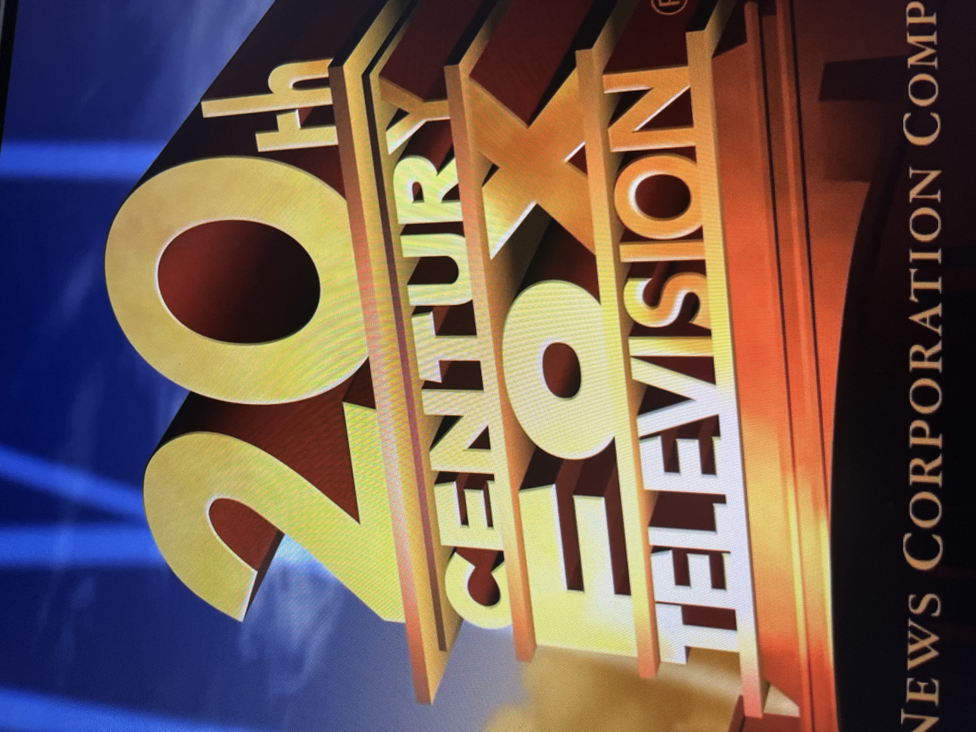 The image is a close-up, low-angle shot of the 20th Century Fox Television logo, rendered in a three-dimensional, metallic gold finish with a reddish-brown gradient on the edges. The logo is presented in a dramatic, illuminated style, with light beams originating from the left and casting shadows. The text "20th CENTURY FOX TELEVISION" is clearly visible, with the "20th" being the largest element on the left, followed by "CENTURY" stacked vertically, and then "FOX" and "TELEVISION" also stacked. To the right of "TELEVISION," the text "NEWS CORPORATION COMP" is visible in a vertical orientation, appearing in a lighter, more subtle font against a dark background. The overall impression is one of grandeur and a cinematic opening sequence. The image appears to be taken indoors, possibly in a studio or on a screen, given the artificial lighting and the nature of the logo display. The background is a deep blue, with subtle streaks of light suggesting a starfield or a dynamic visual effect. There are no people or animals visible in the image. The time of day and weather are not discernible from the image.