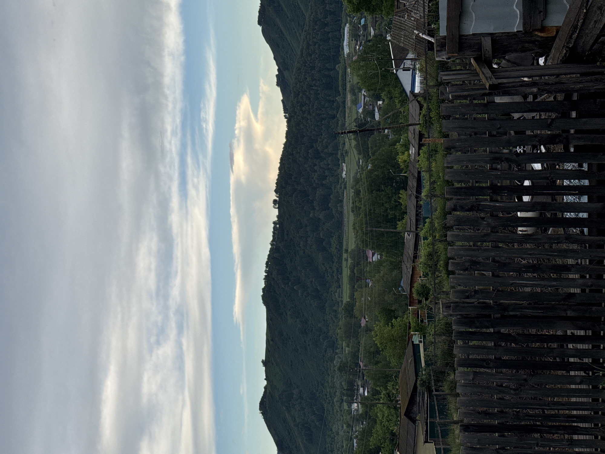 The image captures a view of the village of Soloneshnoye, Russia, overlooking a green, tree-covered mountain range under a cloudy sky. In the foreground, a dark, weathered wooden fence stands prominently. Beyond the fence, the village is visible with scattered houses and structures nestled among lush green trees. Power lines and poles are also present in the middle ground, crisscrossing the landscape. The sky is a mix of blue and white clouds, with a large, illuminated cloud formation visible above the mountains. The scene depicts a rural environment with a natural landscape dominating the background and a residential area in the middle and foreground.