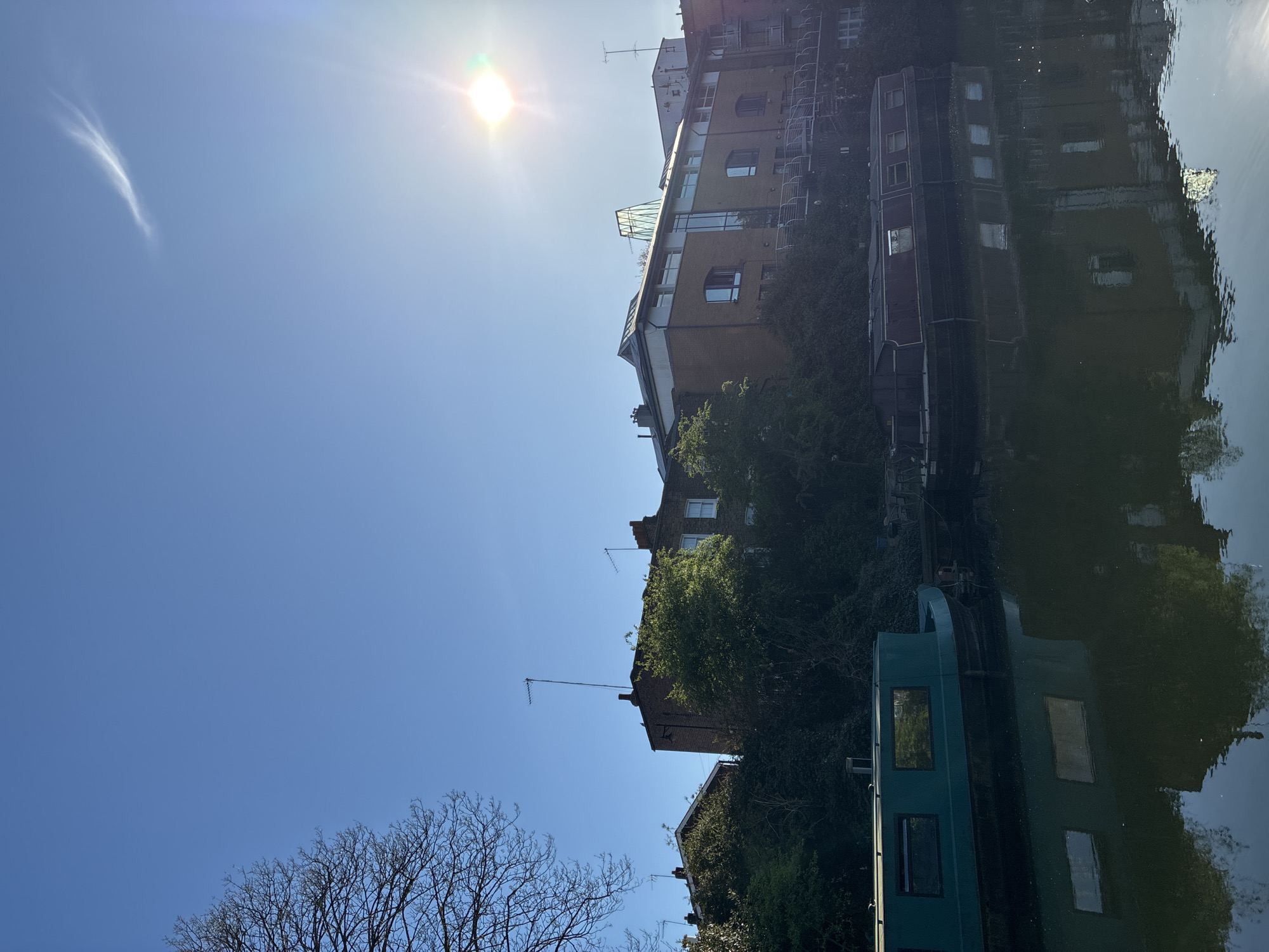 The image depicts a canal in London, United Kingdom, likely Regent's Canal or a similar waterway. The scene is captured on a sunny day, with a bright blue sky and a faint wisp of cloud. Along the canal bank are buildings of varying heights and architectural styles, interspersed with trees and greenery. Several canal boats are moored along the bank, their reflections visible in the water. The water's surface creates a mirror image of the buildings and boats, albeit distorted by the water's movement. The sun's glare is prominent in the sky. A leafless tree is in the foreground.