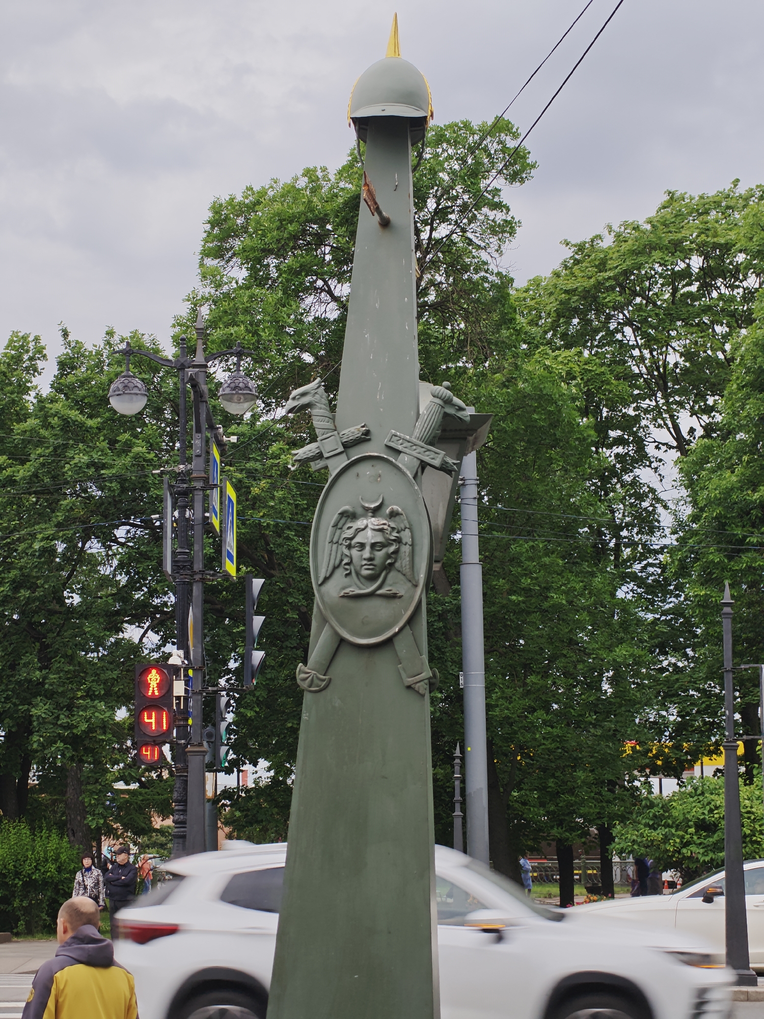 In Saint Petersburg, Russia, a street scene unfolds under an overcast sky, dominated by a large, green metallic lamppost adorned with intricate, stylized military emblems. These emblems feature a shield with a face and winged elements, crossed swords, and a dragon-like figure. The lamppost is topped with a golden finial. To the left, a traffic light displays a red pedestrian signal and numerical readouts of "41" twice. A black lamppost with two globes stands beside it. In the foreground, a white car is captured in motion, with a blurred figure of a person wearing a yellow jacket walking away from the camera. Further into the background, several individuals are visible near a crosswalk, with dense green foliage framing the scene and power lines stretching across the sky.