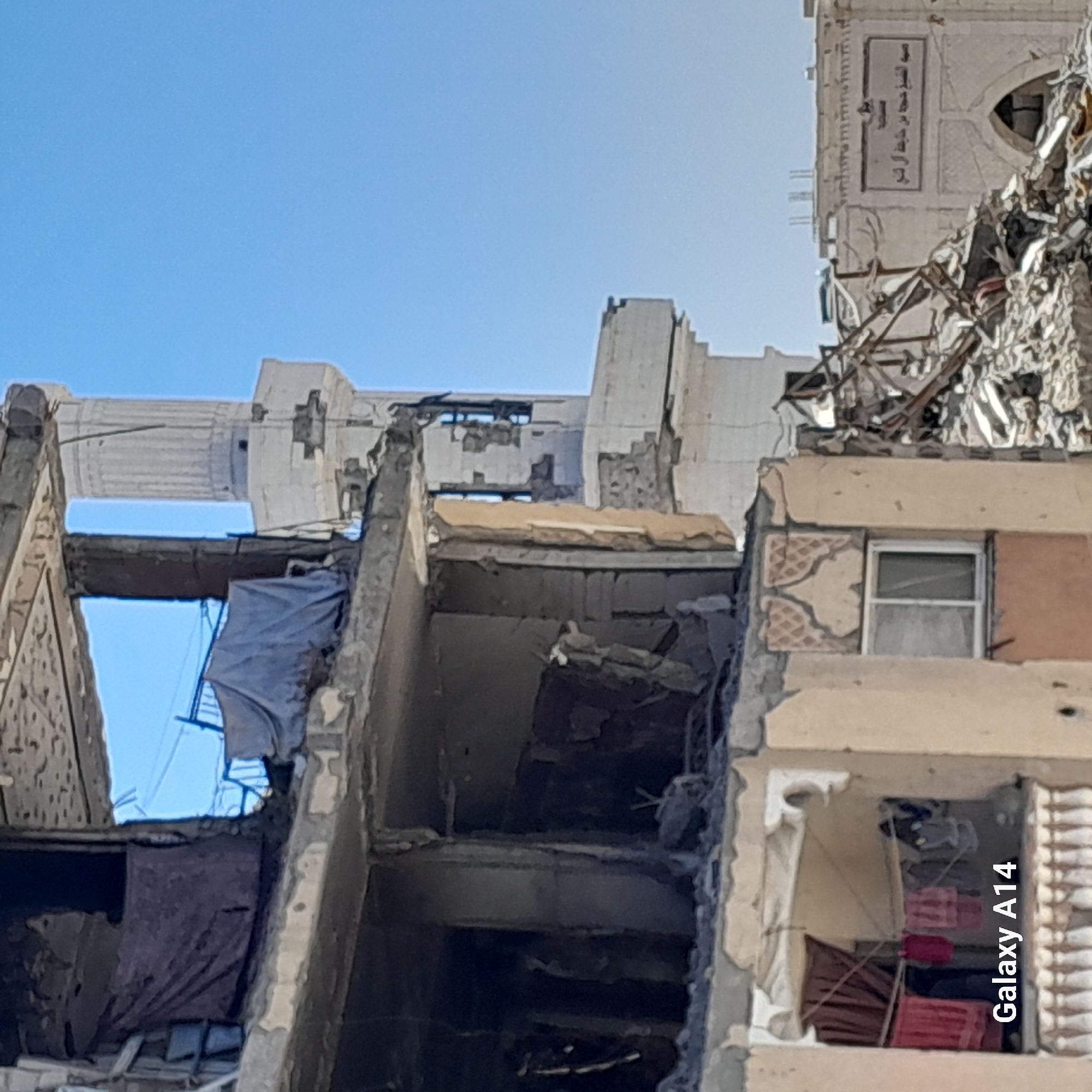 The image shows the aftermath of an airstrike in the city of Khan Yunis, Palestinian Territories. A severely damaged multi-story building is visible in the foreground, with exposed rebar and collapsed concrete. Laundry is hanging on lines within one of the intact sections of the building. In the background, a minaret of a mosque is partially visible, along with other damaged structures and debris. The sky is clear and blue. The image is tagged with "Galaxy A14".