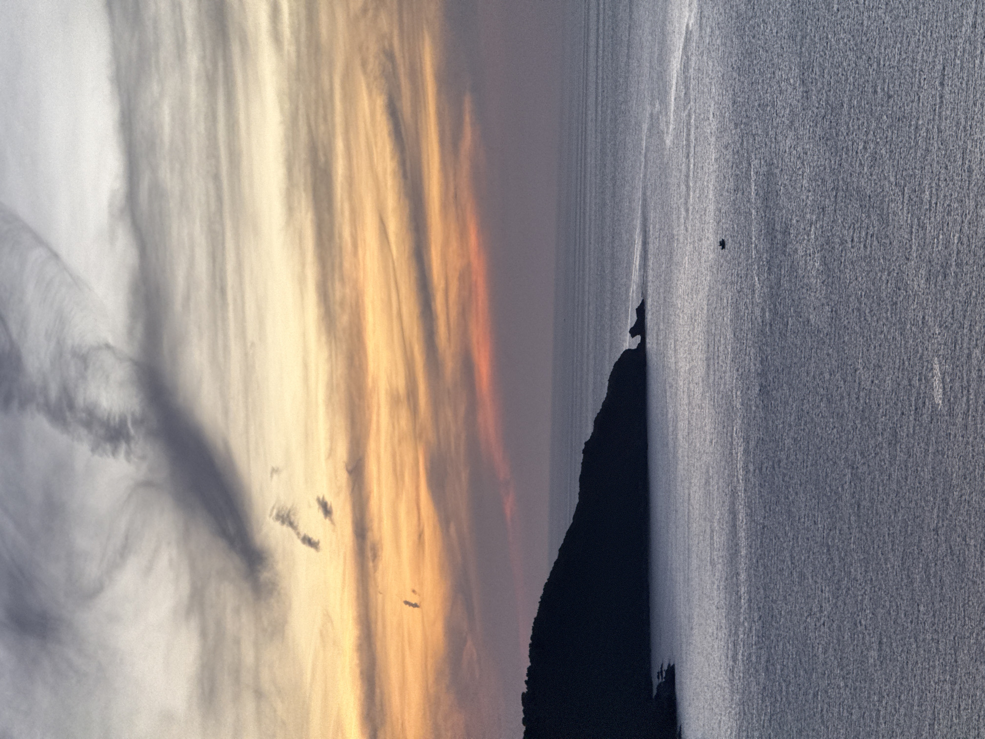 An aerial, wide-angle shot captures a coastal landscape at sunset, likely from Son Tra Peninsula in Da Nang, Vietnam. The sky is a gradient of warm oranges and yellows near the horizon, transitioning into muted purples and grays higher up, with wispy clouds stretching across. A dark, silhouetted headland juts into the calm, silvery sea on the left side of the frame, with the faint outline of smaller rocks visible at its base. A lone, small boat is positioned in the middle of the expansive ocean, adding a sense of scale. The water is largely still, with subtle ripples reflecting the twilight sky. The overall atmosphere is serene and tranquil, emphasizing the natural beauty of the coastline at dusk.