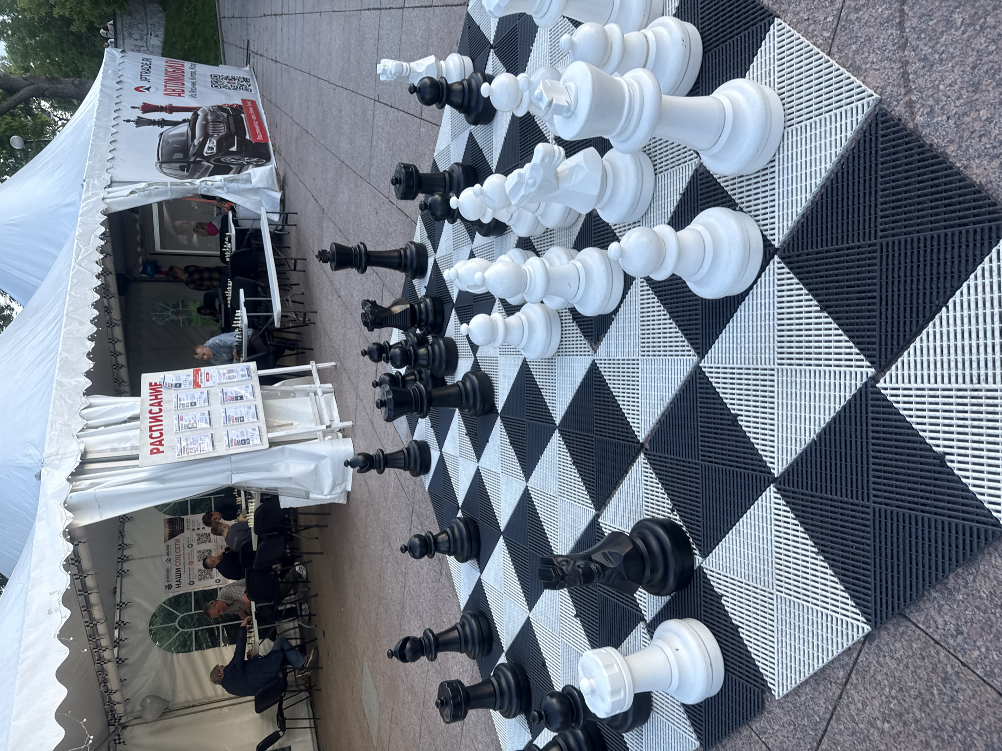 An outdoor chess event is taking place under a white tent in Vladivostok, Russia. A large, portable chessboard is set up in the foreground with oversized black and white chess pieces. In the background, several people are seated at tables playing chess, with more tables and chairs visible. A sign in Russian, likely displaying a schedule, is propped up on an easel. Banners with advertisements are displayed on the tent walls, one featuring a car and the text "JPTRADER.RU АВТОМОБИЛИ". The ground appears to be paved with stone tiles.