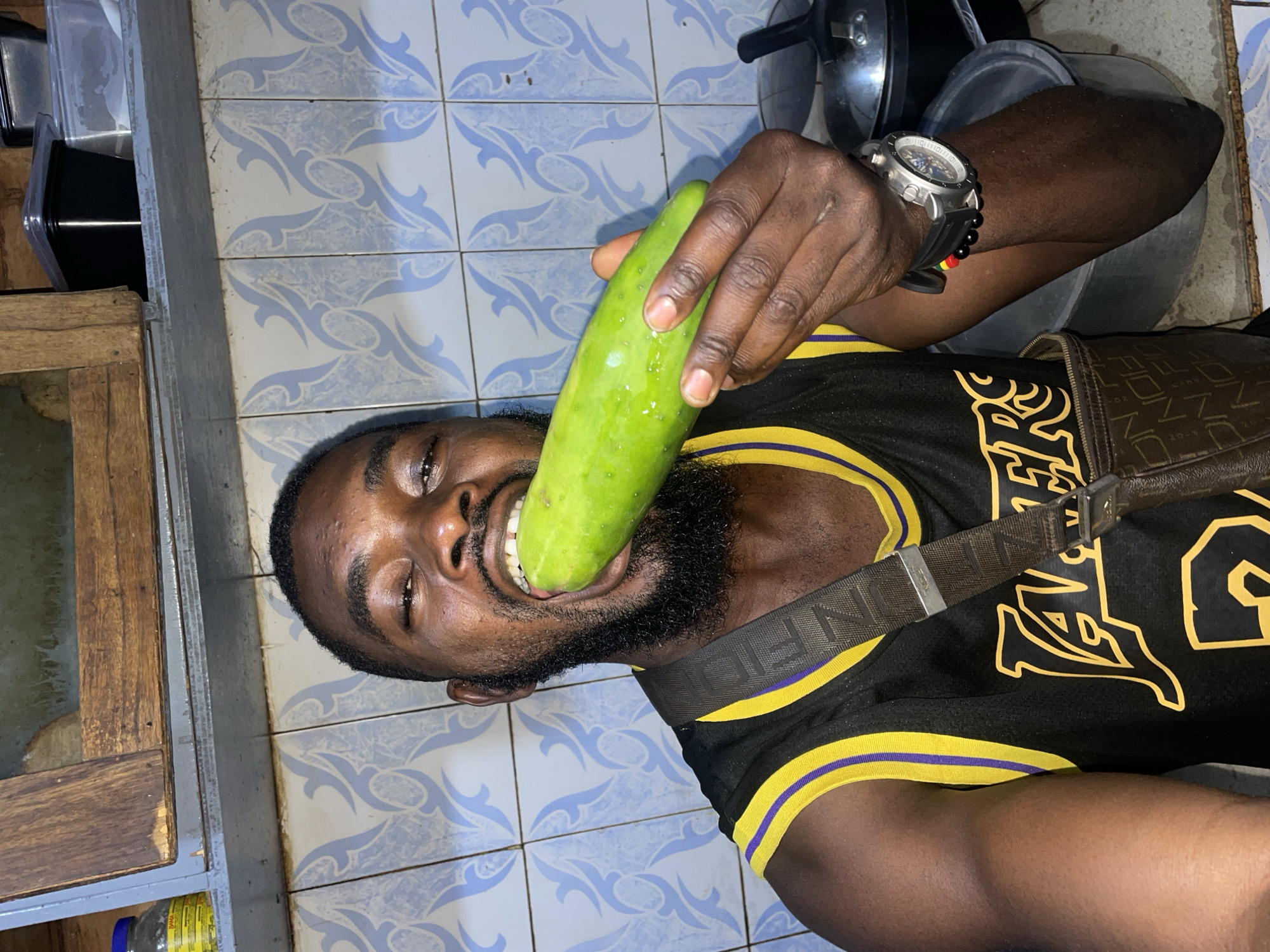 A person is holding a cucumber to their mouth. They have dark skin, a beard, and are wearing a black and yellow basketball jersey with a shoulder bag. The background shows blue and white patterned tiles, a wooden cabinet, and some kitchen utensils. The picture seems to be taken indoors, potentially in the city of Jos, Nigeria.