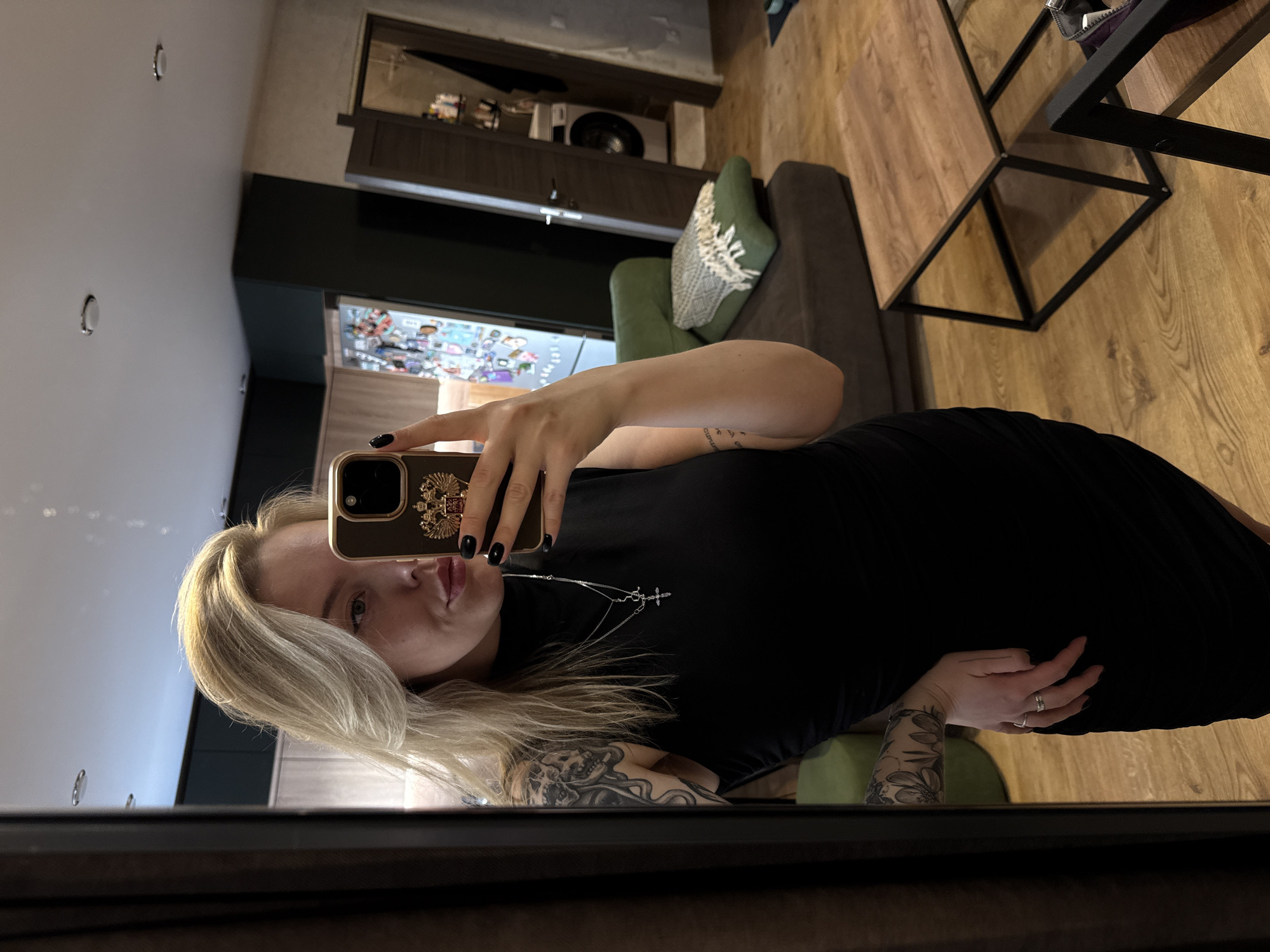 A woman is taking a selfie in a mirror. She has blonde hair and is wearing a black dress. Her phone has a case with a golden emblem on it. She has tattoos on her arms and a necklace with a cross pendant. In the background, there's a glimpse of an interior with a couch, a table, and a cabinet. A collection of objects is visible behind a glass door of the cabinet. Based on the coat of arms on the phone, it is likely that the location is in Russia. Given other visual cues, the location may be the city of Krasnoyarsk, Russia.