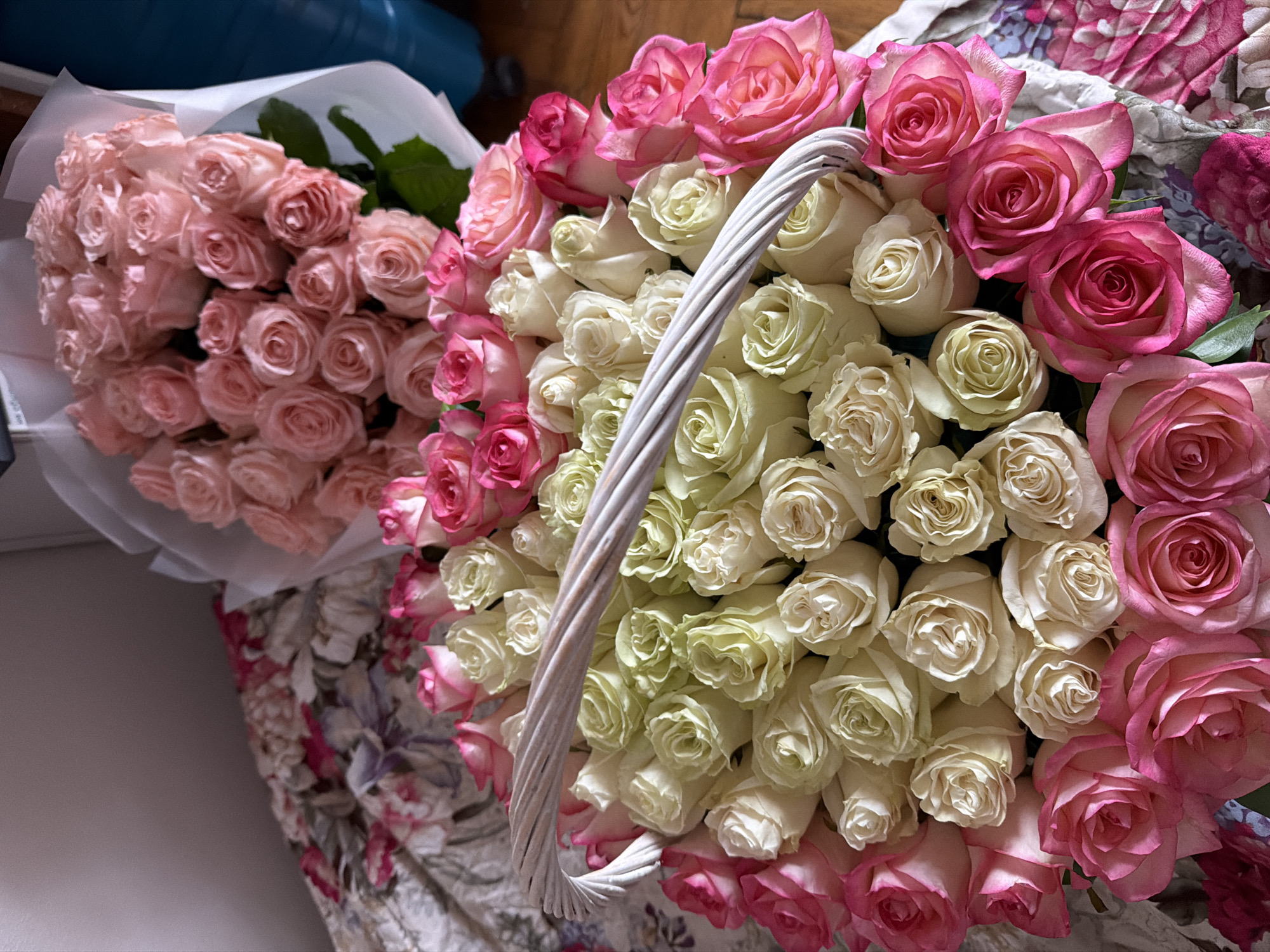 The image displays two large arrangements of fresh roses, set indoors in Moscow, Russia. On the left, a bouquet wrapped in white paper showcases numerous light pink roses, densely clustered and slightly out of focus. In the foreground on the right, a white woven basket is filled with a vibrant assortment of roses. The central area of the basket is packed with white or cream-colored roses, while the outer edges are bordered with bright fuchsia and deep pink roses, creating a striking contrast. The white, braided handle of the basket arches over the center of the floral arrangement.
In the background, a floral-patterned fabric, likely a quilt or blanket featuring shades of pink, purple, and white, is visible on the lower right. A blurry dark blue object is partially seen in the upper left corner, and a plain, light-colored surface occupies the lower left background. The lighting is soft, indicating an indoor setting. No people are visible, and there is no discernible text in the image. The scene suggests a celebration, a special occasion, or a gift of flowers.