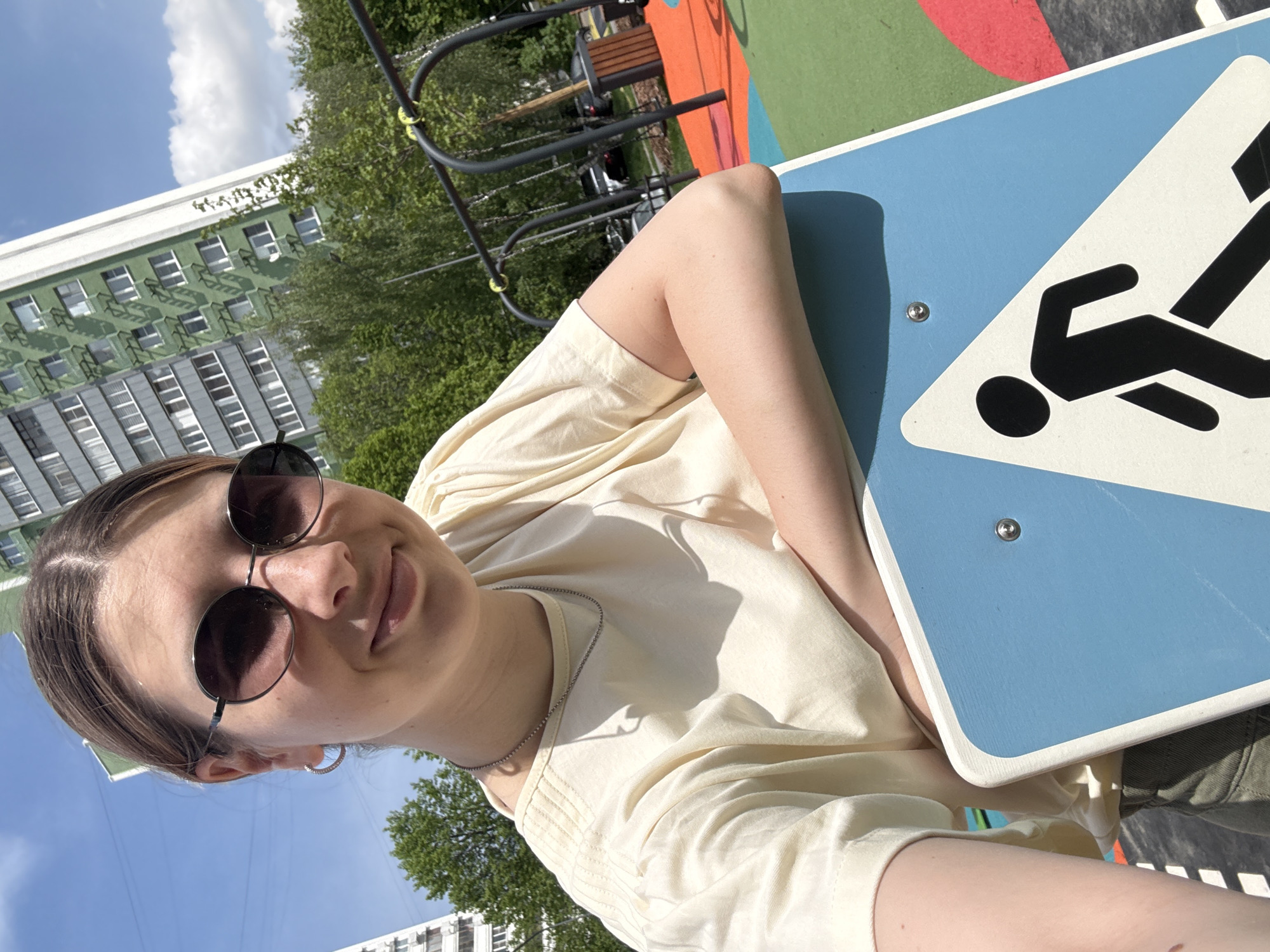 A young woman in sunglasses is taking a selfie outdoors. She is wearing a light-yellow t-shirt and a silver chain necklace. A blue and white pedestrian crossing sign is to her right. The location appears to be a playground, with a portion of a swing set visible. In the background, a building with multiple windows is visible, along with trees and a blue sky. The picture was taken on a bright sunny day. It is believed that the location is in the city of Moscow, Russia.