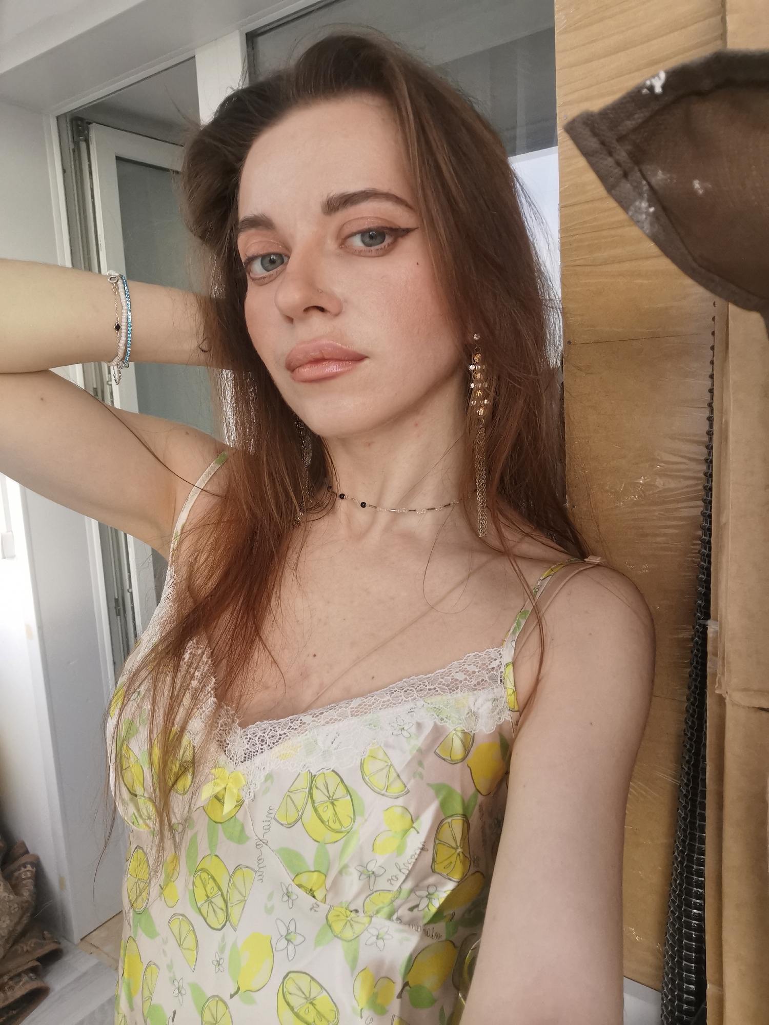 A young woman with long, flowing brown hair poses for a headshot-style portrait, looking directly at the camera with a neutral expression. Her light blue-grey eyes are accentuated with winged eyeliner, and her lips are painted with glossy nude-pink lipstick. She has a fair complexion.
She is wearing a light-colored camisole or slip adorned with a pattern of yellow lemons, green leaves, and white lemon blossoms. The neckline features a white lace trim, and faint, illegible script is subtly visible on the fabric. On her right wrist, she wears multiple bracelets, including one with white or pink beads, another with blue rhinestones, and a silver chain. Two delicate necklaces are visible around her neck: a dark beaded choker and a thin silver chain. A long, dangling earring with golden or amber-colored beads and chains hangs from her left ear. Her left arm is raised, with her hand positioned near her head.
The background on the left reveals a white window or door frame, suggesting an indoor or sheltered outdoor setting. On the right, there is a stack of brown cardboard boxes, with some dark, mesh-like material visible between them. The lighting in the image is bright, indicating daylight.