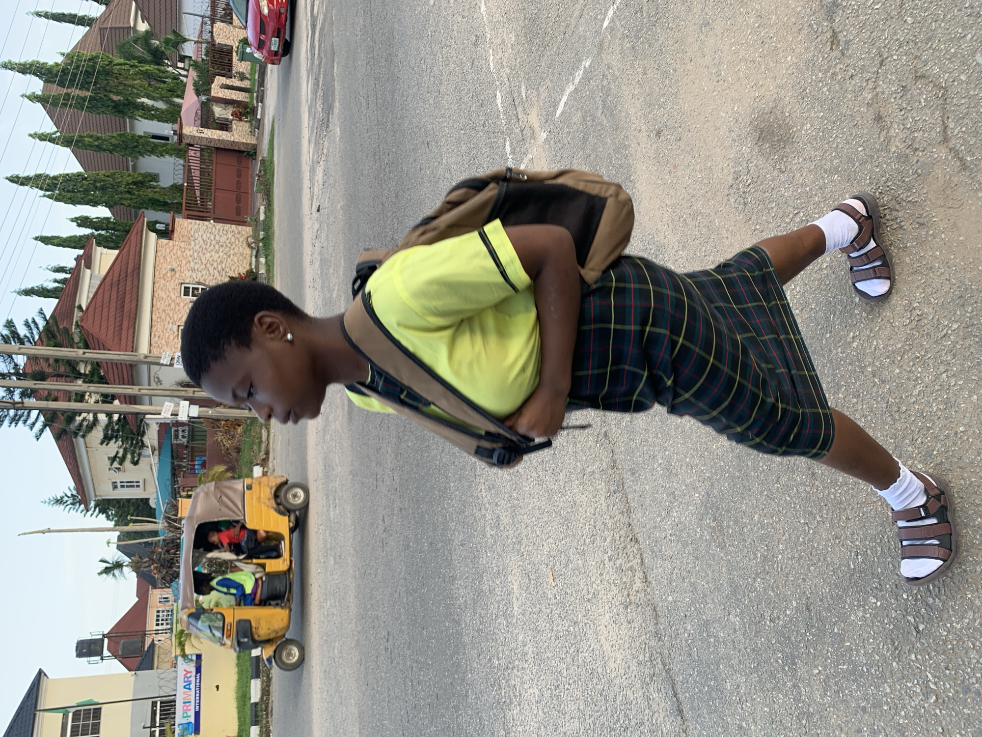 The image captures a side-view of a young Black female walking on a paved road in Abuja, Nigeria. She is wearing a lime green short-sleeved shirt with black trim, a dark plaid skirt, white socks, and brown sandals. A beige backpack is slung over her shoulders. The background shows a residential area with modern houses, some featuring red-tiled roofs and tall green trees. A yellow auto-rickshaw, also known as a 'keke', is visible in the distance, carrying passengers. A red car is parked further down the road on the right. The sky is overcast. The scene appears to be a typical street view in a suburban area of Abuja.