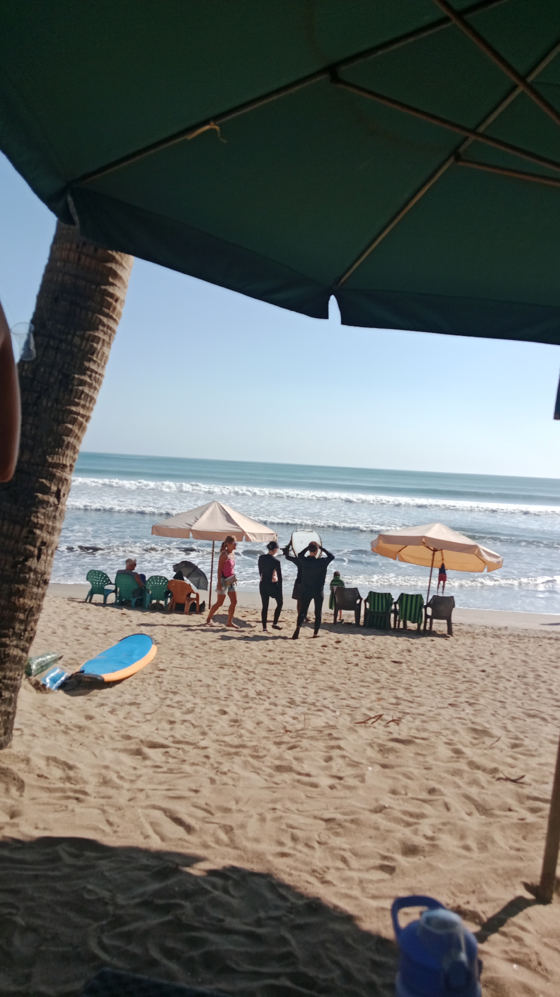 The image depicts a beach scene in Kuta, Indonesia, with the ocean in the background and sand in the foreground. Several beach umbrellas are set up, providing shade. A group of people are visible, some sitting on chairs and others standing. Two individuals in black wetsuits appear to be posing with surfboards. A blue and orange surfboard lies on the sand. A coconut palm tree is present on the left side of the image. The sky is clear and blue.