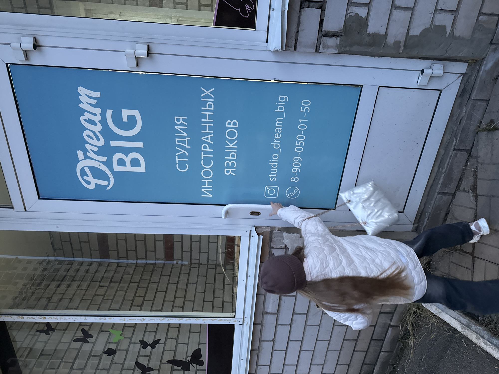 A woman with long brown hair is opening the door to a business identified as "Dream BIG" in Izhevsk, Russia. She is wearing a dark-colored beanie, a white puffy jacket, dark pants, and white shoes. She appears to be holding a white bag with a rope. The door is glass with a light blue sign displaying "Dream BIG" in English and Cyrillic script, along with an Instagram handle "studio_dream_big" and a telephone number "8-909-050-01-50". The window to the left of the door is decorated with black butterfly stickers. The building is brick. The sidewalk is paved.
