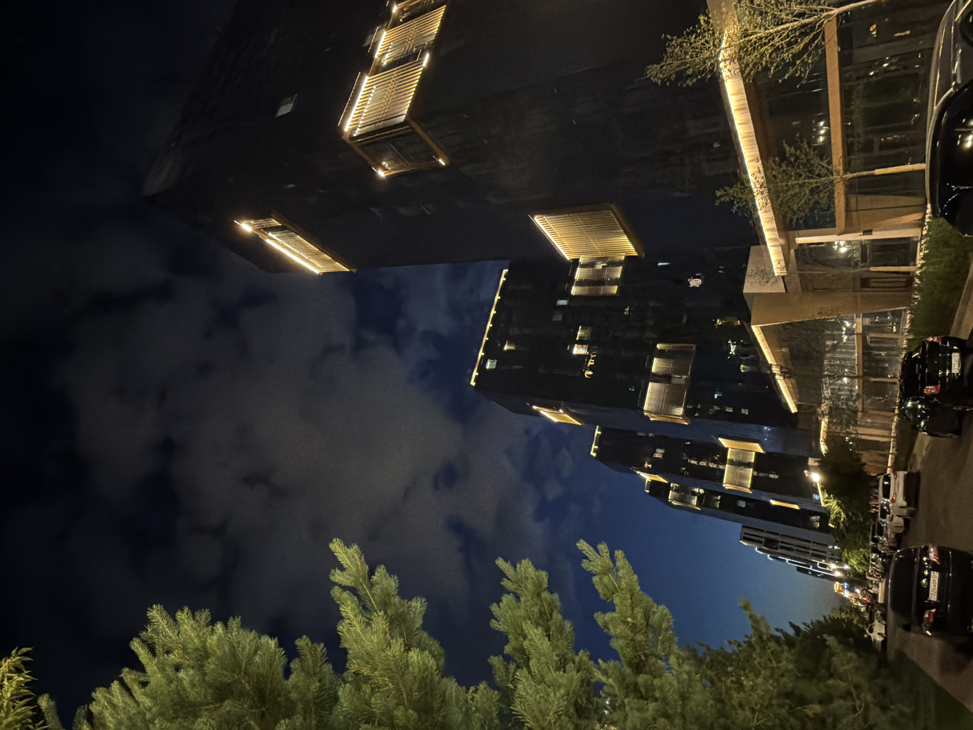 The image depicts a contemporary cityscape at night in Astana, Kazakhstan, specifically along the Avenue of the First President. The scene is dominated by a row of modern, dark-colored buildings with striking architectural features, characterized by illuminated rectangular window frames and balconies that glow with warm, yellow light. A lush green coniferous tree stands prominently in the foreground on the left, its branches silhouetted against the darkening sky. The sky itself is a deep blue with scattered, wispy clouds, suggesting a twilight or early night setting. A street lined with parked cars stretches into the distance, with vehicles of various colors, including black and white sedans and SUVs, visible. The buildings appear to be a mix of residential and commercial spaces, with a ground-level section featuring large glass windows and a warm, inviting light emanating from within. The overall impression is one of urban sophistication and modern design, illuminated by artificial light against the natural darkness of the night.