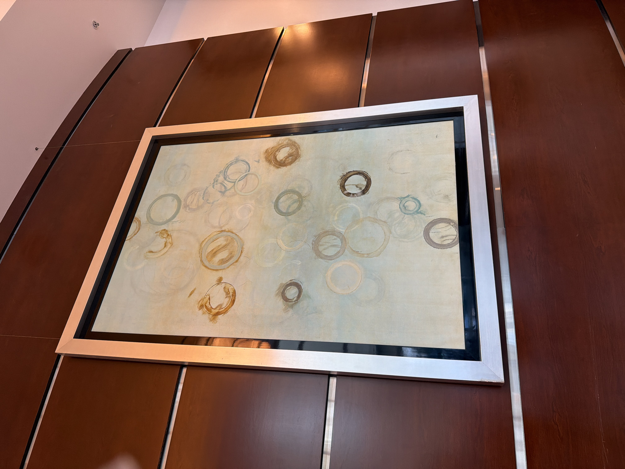 The image shows a framed abstract painting hanging on a wood-paneled wall. The painting features circular shapes in varying shades of beige, brown, and light blue on a pale background. The frame is wide and appears to be silver or a metallic light color with a black inner border. The wall is composed of horizontal wooden panels with thin silver trim between them. The lighting suggests an indoor environment, possibly a lobby or a gallery. There are no people or specific landmarks visible that would indicate a location in San Diego, United States of America.