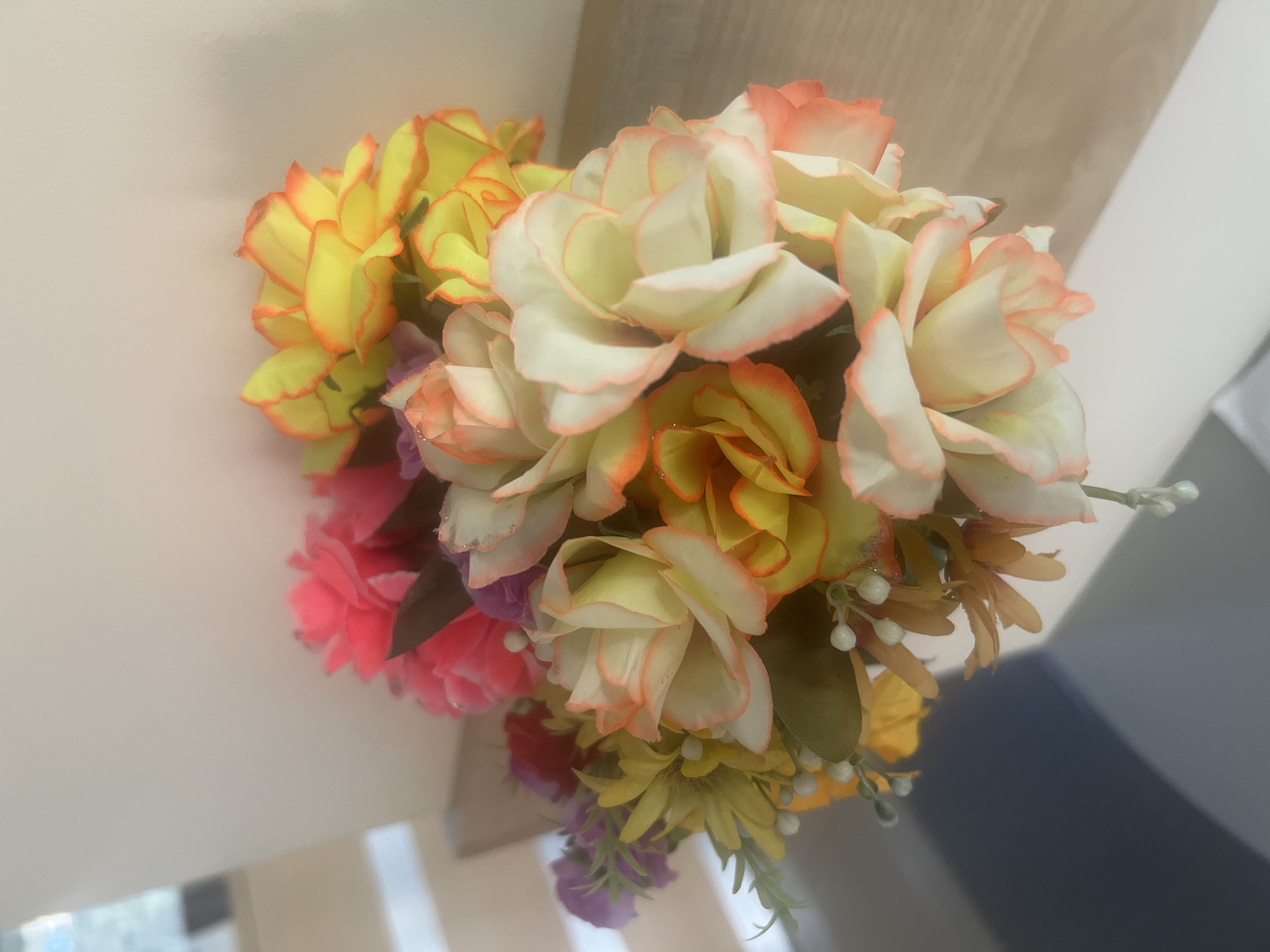 A close-up, slightly out-of-focus shot shows a vibrant bouquet of artificial flowers. The flowers are in various stages of bloom, with prominent shades of yellow, orange, white, pink, and purple. Some of the flowers have delicate pink or orange-tinged edges. Small white buds and green foliage are interspersed throughout the arrangement. The bouquet is positioned on a light-colored, wooden surface, likely a shelf or table, with a light-colored wall in the background. A subtle shadow of the bouquet is cast on the lower left of the frame, suggesting a light source from the right. The environment appears to be indoors, possibly a home or office setting. There are no people or animals present in the image. The specific location within the city of Magas, Russia, cannot be determined from the visual information provided.