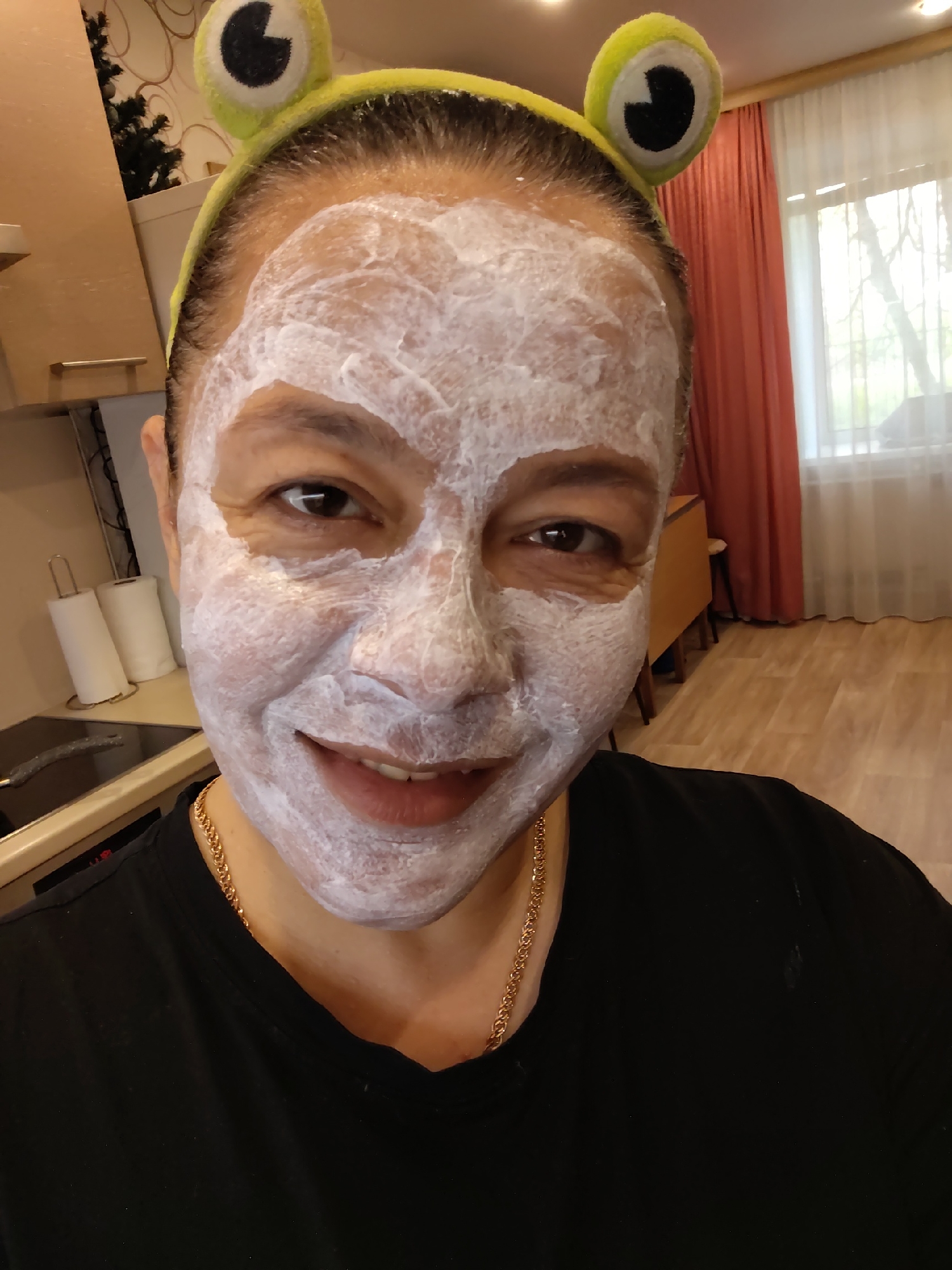 A close-up selfie shows a middle-aged woman wearing a black t-shirt and a yellow headband with protruding cartoon frog eyes. Her face is covered in a thick, white facial mask. She is smiling slightly, exposing her teeth. A gold chain is visible around her neck.
The background suggests a domestic interior. A kitchen area is partially visible on the left, featuring light-colored cabinets, a black stovetop, and a roll of paper towels. To the right, a window is covered with sheer white curtains and red drapes. An area with wooden flooring is visible beyond the drapes. The location is Unknown, Unknown.