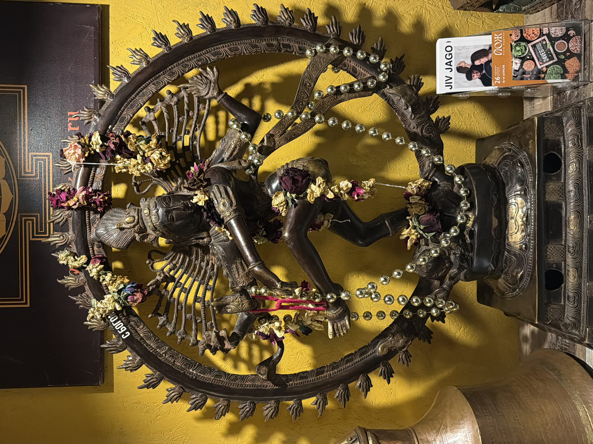 The image shows a statue of Nataraja, the dancing form of Shiva, adorned with garlands of dried flowers and a string of golden beads. The statue is mounted on a wall, likely indoors. To the left is a dark-colored object with golden details, and to the upper right, there is a magazine or booklet titled "JIV JAGO" with images of people and food. The background wall is painted in a mustard yellow color. The statue appears to be located in a place with religious significance. There are no people present in the image. Based on the magazine title in Russian "ЙОЖ Йога журнал", this picture was taken in Moscow, Russia.