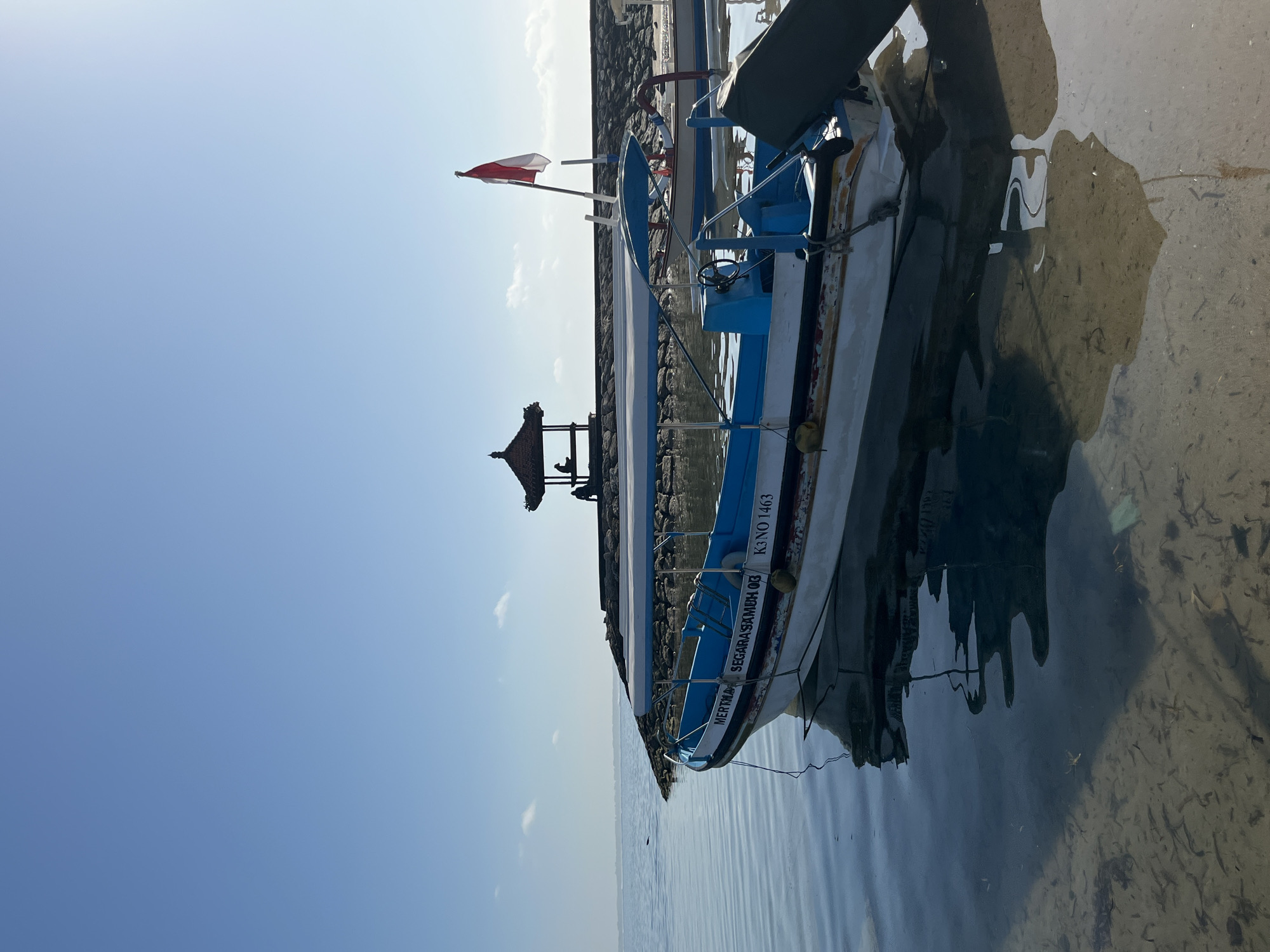 In Kuta Selatan, Indonesia, a clear blue sky stretches over a tranquil body of water. In the foreground, a white and blue boat is moored, its reflection rippling on the glassy surface. The boat is equipped with a canopy and appears to be a small passenger vessel, with the text "MERTEGA SEGARASAMEH OG" and "K3 NO 1463" visible on its side. Further in the background, a stone jetty extends into the water, topped by a traditional Balinese gazebo. Two figures are seated within the gazebo, observing the serene seascape. The water is shallow enough to reveal the sandy bottom and sparse vegetation, indicating a calm and clear environment. The scene suggests a peaceful morning in a coastal area, possibly a harbor or a quiet beach.