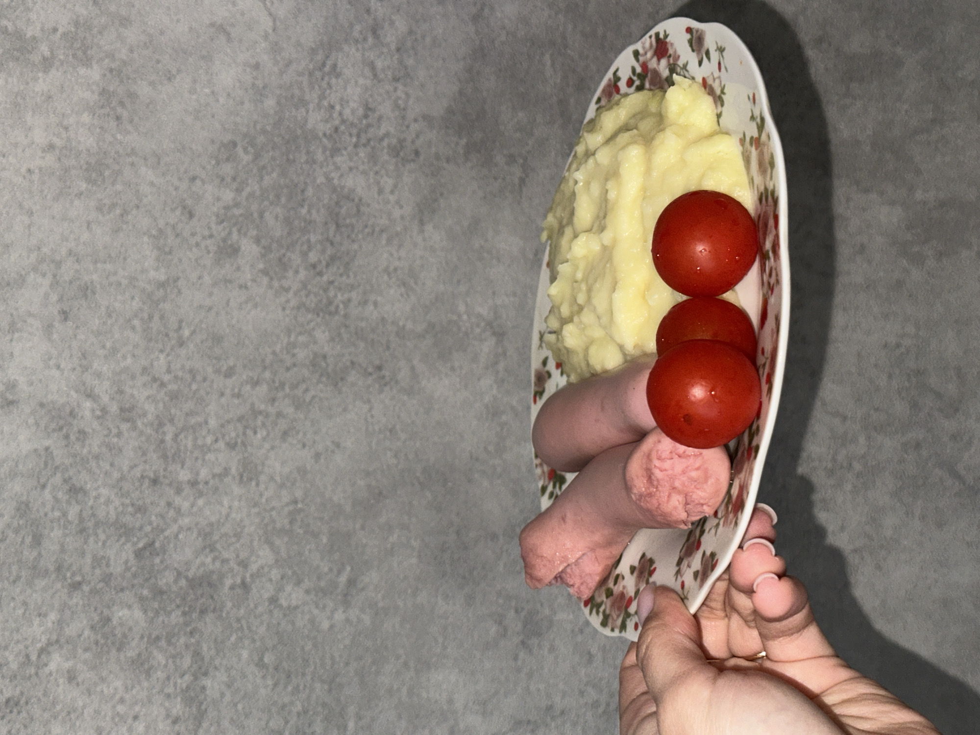 A close-up shot shows a plate of food held in a person's hand. The plate contains mashed potatoes, three red cherry tomatoes stacked atop each other, and two sausages. The plate is white with a floral pattern along the edges. The hand holding the plate is light-skinned and appears to be manicured. The background is a textured gray surface.
The location is not discernible from the image. There is no indication that the photo was taken in Chaikovsky, Russia.