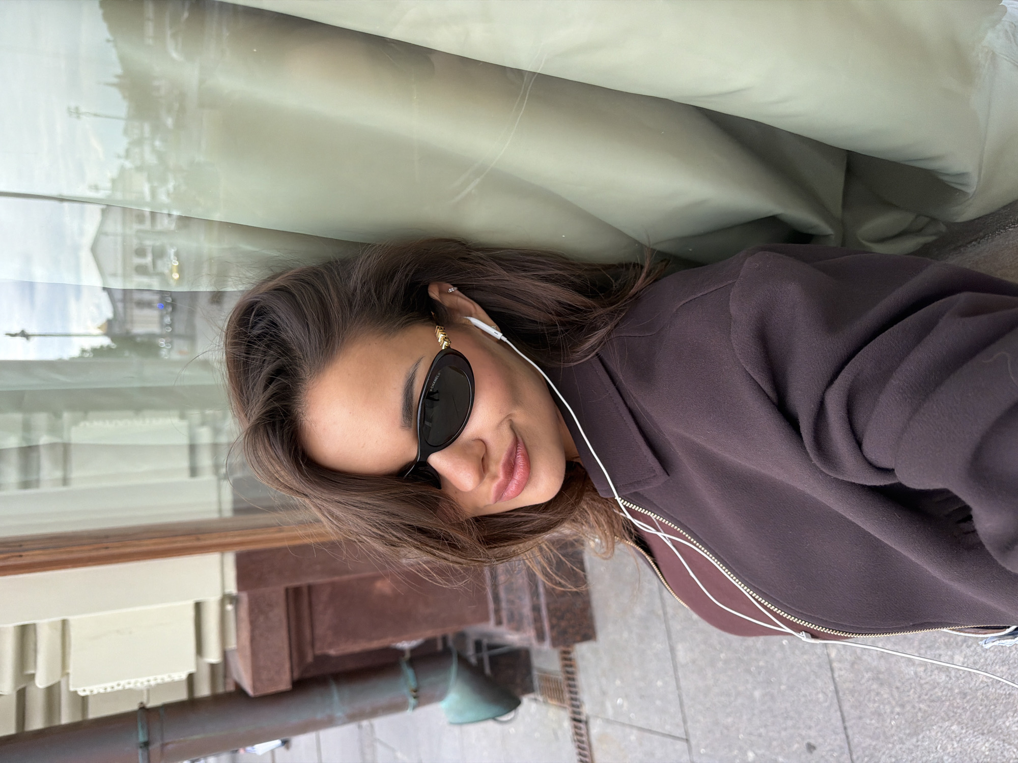 The image, presented with a 90-degree clockwise rotation, features a young woman leaning or lying against a reflective surface in an urban setting. Her head is positioned towards the left side of the frame, and her body extends towards the right.
The woman has long, dark brown hair and is wearing black oval-shaped sunglasses, which clearly display the "CHANEL®" brand name on the arm. White wired earphones, with "SONY" visible on one earpiece, are plugged into her ears, and the wires trail down her dark brown zip-up jacket. She has a relaxed, slight smile.
Above her head (which would be the upper part of the image if oriented upright), a blurry reflection of city buildings and structures is visible on the surface she's leaning against, indicating an outdoor, urban environment. The reflections suggest daytime, possibly under an overcast sky, with soft lighting.
Below her (which would be the lower part of the image if oriented upright), a grey paved surface, consistent with a sidewalk, is seen. Adjacent to this, there are dark reddish-brown stone architectural elements and a dark, possibly metallic, pipe-like structure. These details reinforce the outdoor, city location, consistent with Moscow. The woman appears to be posing casually for the camera, possibly listening to audio.