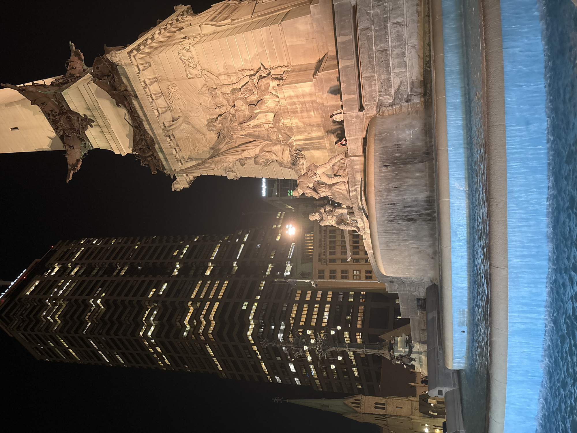 This nighttime photograph captures a portion of the Soldiers and Sailors Monument in Indianapolis, Indiana, USA, a memorial in the city's downtown area. The monument's ornate stonework, including sculpted figures and reliefs, is visible, illuminated by artificial lighting. To the right of the frame, water from the monument's fountain is shown. In the background, the cityscape includes the brick-clad skyscraper at 101 West Washington Street. A steeple from nearby Christ Church Cathedral can be seen at the bottom of the image. The scene is dark with the artificial lights causing the most illumination in the image.