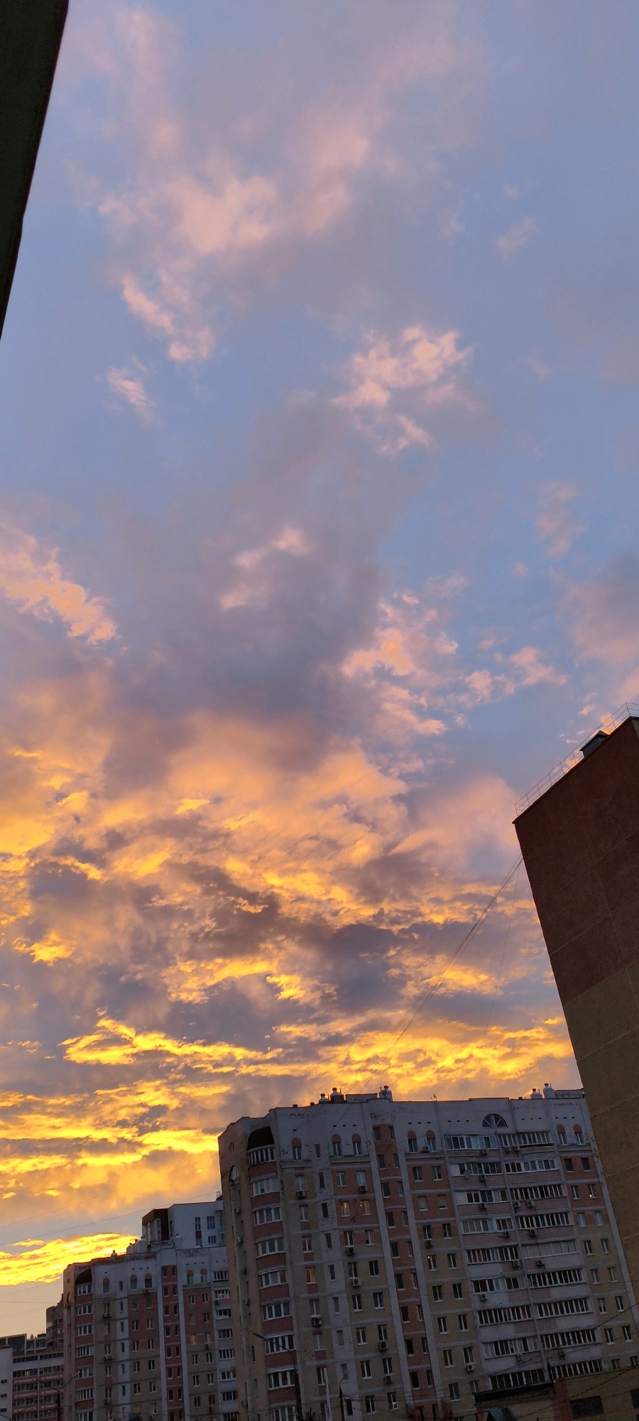 A vertical shot captures a cityscape at dusk. The sky is a dramatic canvas of clouds, with hues of orange and yellow dominating the lower portion, suggesting the setting sun. Above, the sky transitions to a soft blue, punctuated by lighter pinkish clouds.
Dominating the lower and right portions of the image are the façades of apartment buildings. The buildings are multi-story structures with a visible pattern of windows and balconies. One building on the right is primarily brick-red, while the buildings in the center and left are a lighter color, possibly beige or light gray, with accents of pink and white. Some windows appear lit, indicating occupancy. Utility wires are visible stretching across the frame between buildings. A dark, triangular shape, possibly a rooftop or structural element of another building, juts in from the top left corner. No people are visible in the scene.