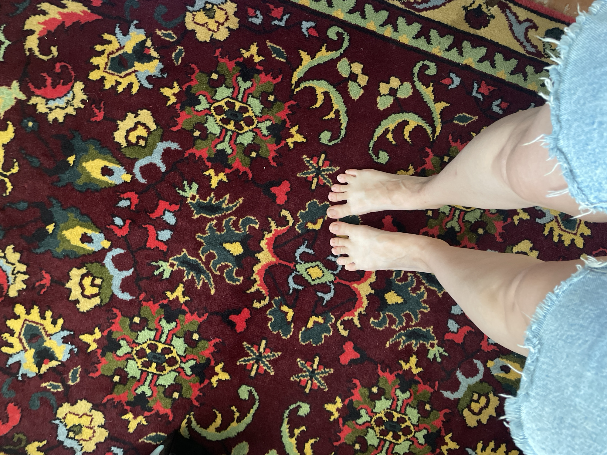 The image shows a person's bare feet and lower legs standing on a burgundy rug with a floral pattern. The rug features intricate designs in yellow, green, red, and blue. The person is wearing light blue denim shorts with frayed edges. The scene is photographed from a high angle, looking down at the person's feet and the rug. The location is not specified in the image, and there is no visual information to indicate it is in Kopeysk, Russia.