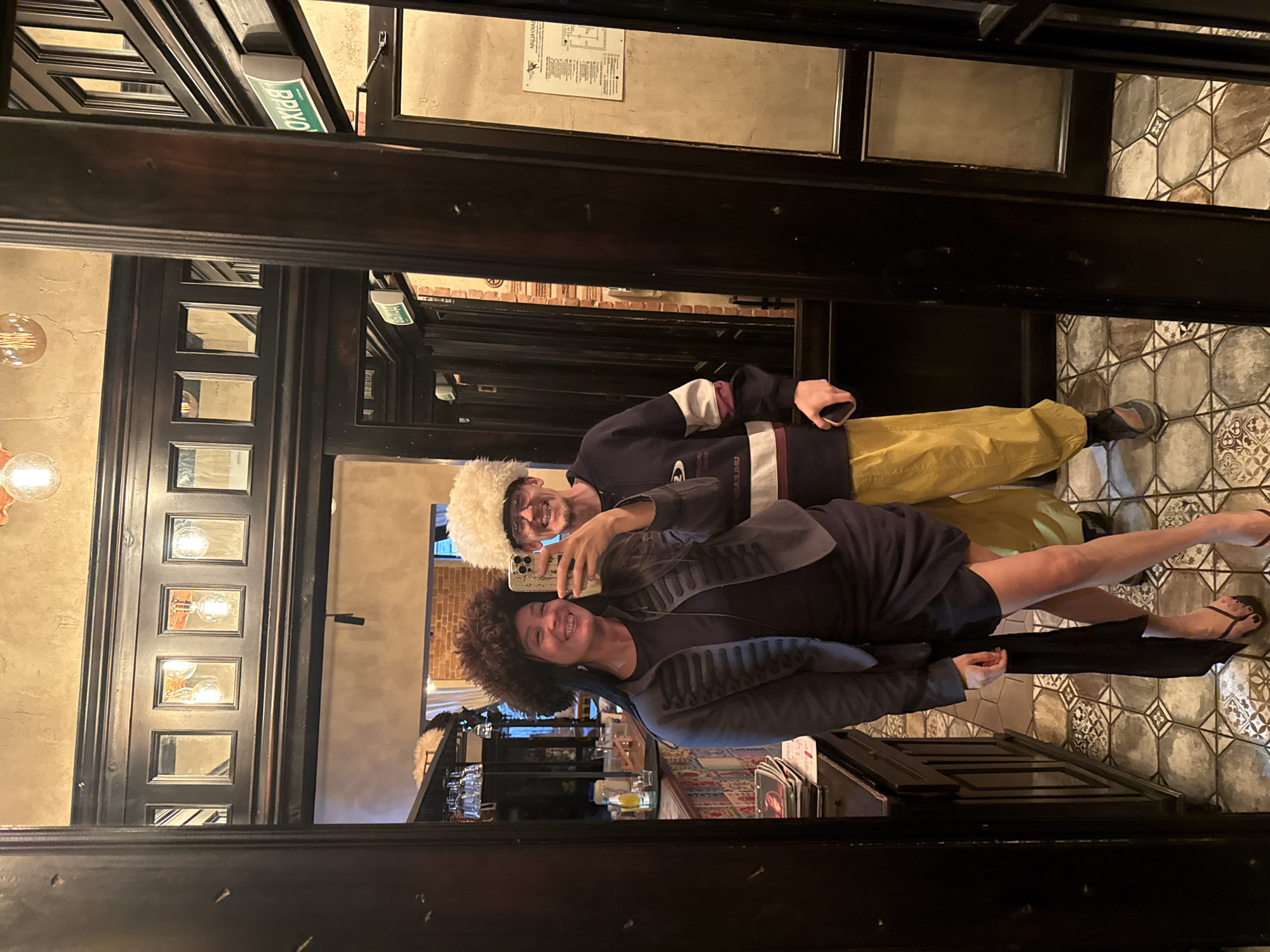 In Saint Petersburg, Russia, two individuals are reflected in a mirror, posing for a selfie. The woman in the foreground, on the left, wears a dark blue dress and a dark jacket adorned with decorative buttons. She has a large, furry brown hat on her head. The man, on the right, is wearing a dark blue sweatshirt with white and maroon stripes on the sleeves and a light-colored, fluffy hat. He also sports glasses and yellow pants. They are standing in an interior space with wooden paneling and tiled floors. In the background, a bar area is visible with shelves of glassware. An illuminated "EXIT" sign in green Cyrillic characters is mounted on the wall, indicating an exit door. The overall lighting suggests an indoor evening setting.