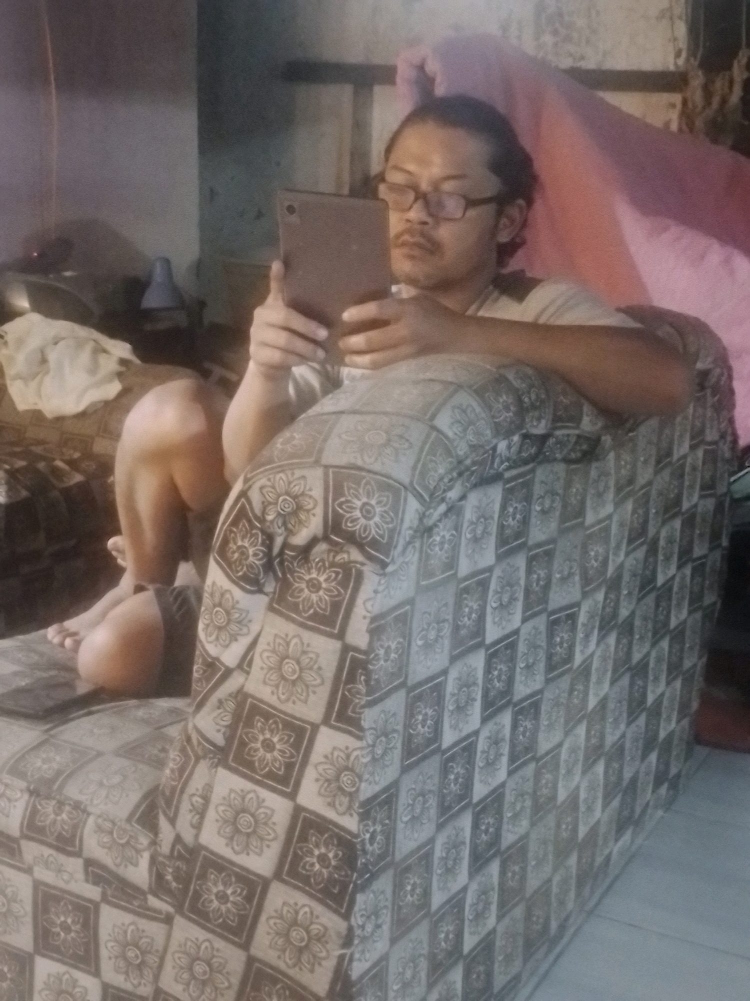 An adult male is seated on a patterned couch, holding a tablet device. He is wearing glasses and a light-colored t-shirt. His legs are crossed, and his right foot is resting on the couch. The couch is upholstered with a repeating floral diamond pattern. In the background, there is a pink blanket draped over something. To the left of the couch, a portion of another couch or furniture is visible, with a white cloth draped on it. A desk lamp is also visible in the background. The scene appears to be indoors, in what seems to be a residential setting.