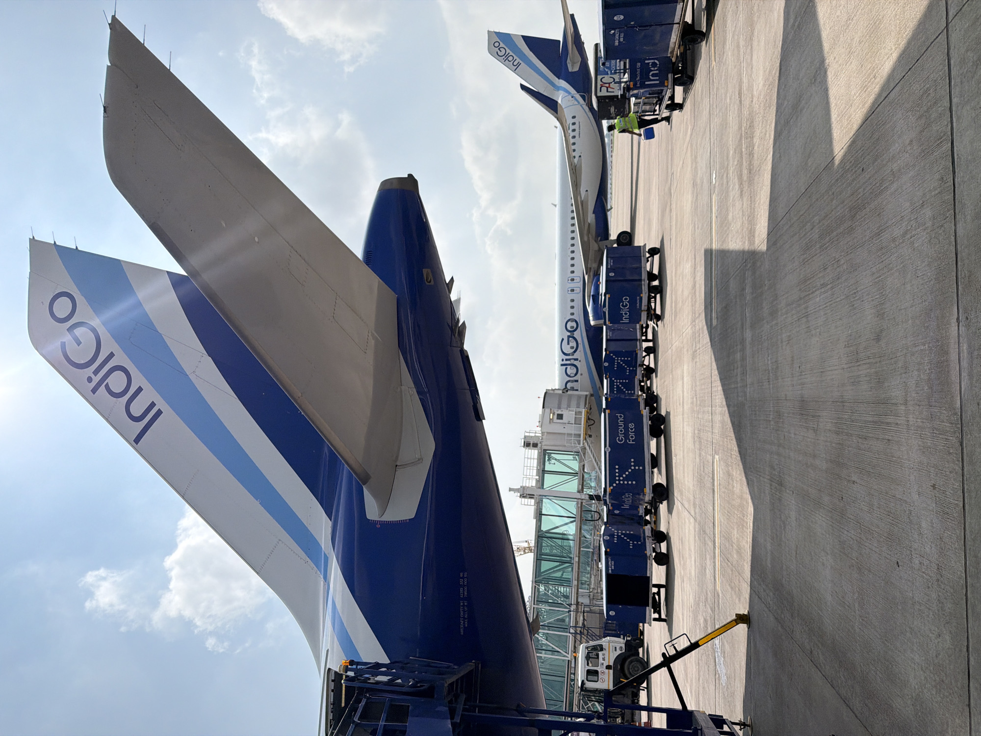 An Indigo Airlines airplane is parked on the tarmac at an airport in Vikram Nagar, India. The tail of the airplane, with the Indigo logo, is visible. Several baggage carts are lined up on the tarmac, some with the Indigo logo and "Ground Force" printed on them. A worker in a yellow vest is walking near the carts. In the background, another Indigo Airlines plane is visible, along with a jet bridge and a ground vehicle. The sky is blue with some clouds.