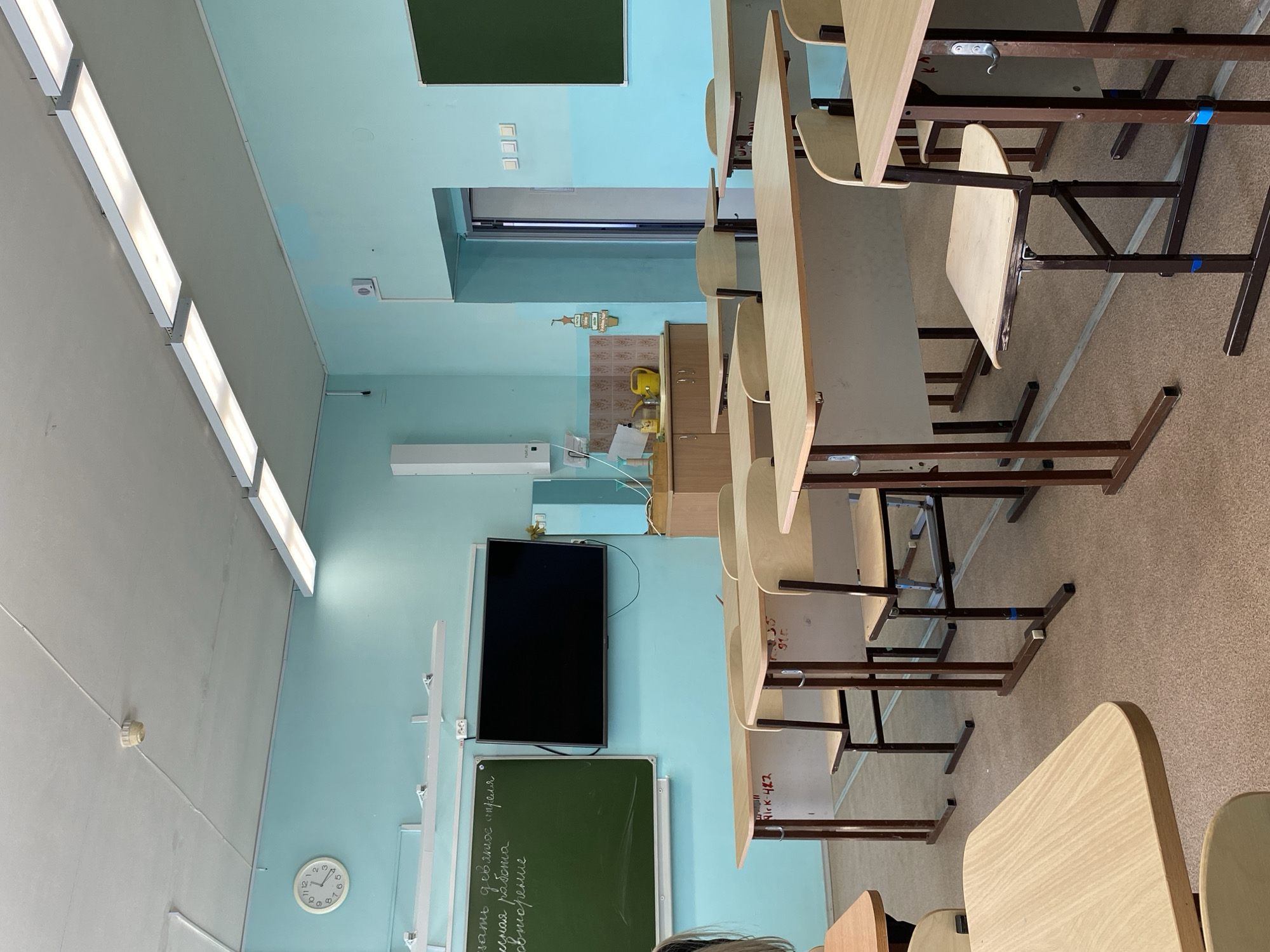 The image shows an empty classroom in Votkinsk, Russia. The room is painted in light blue and white. There are rows of light wooden desks and chairs arranged in neat lines. At the front of the classroom is a black television screen and a green chalkboard with writing on it in Russian. A white clock is visible on the wall. There is a window and some cabinets at the back of the room.